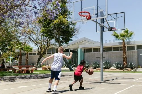 Two children playing basketball on an outdoor court at a school, with trees, plants, and a building in the background.