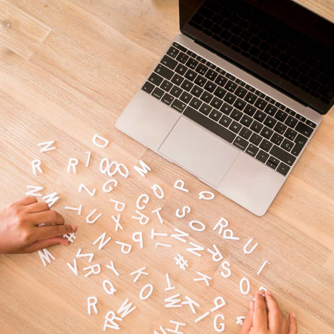 A silver laptop on a wooden desk with scattered white alphabet letter tiles and hands arranging the tiles.