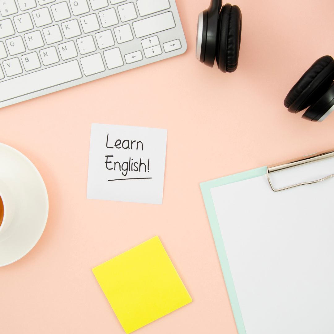 Desk with a white keyboard, black headphones, a cup of tea, yellow sticky note, a clipboard, and a paper with 'Learn English!' written on it.