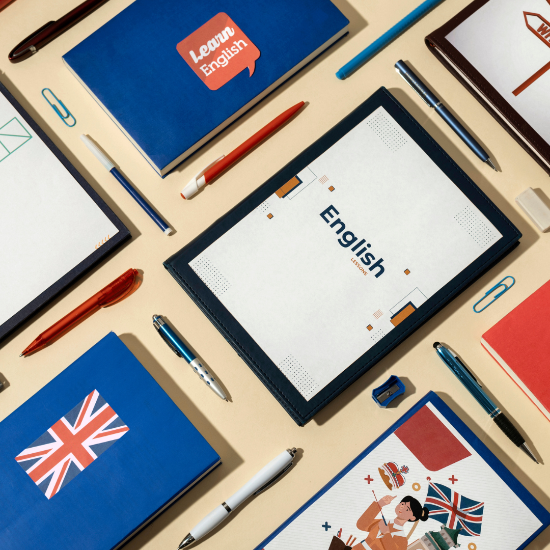 Educational materials for learning English, including notebooks labeled 'Learn English' and 'English lessons,' pens, pencils, paper clips, a notebook with a British flag, and a folder with an illustration of a woman and British symbols, arranged on a beige desk.