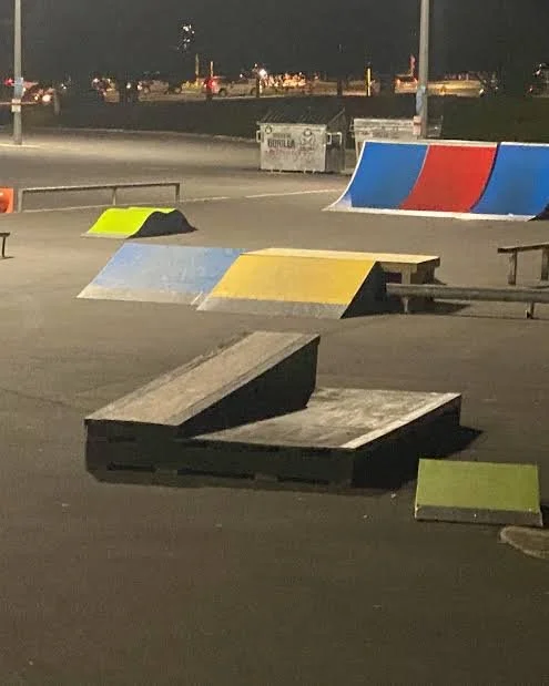Empty skate park at night with ramps and obstacles, including a black ramp, a yellow and blue ramp, and a curved barrier painted in red, white, and blue. Streetlights and a parking lot are visible in the background.