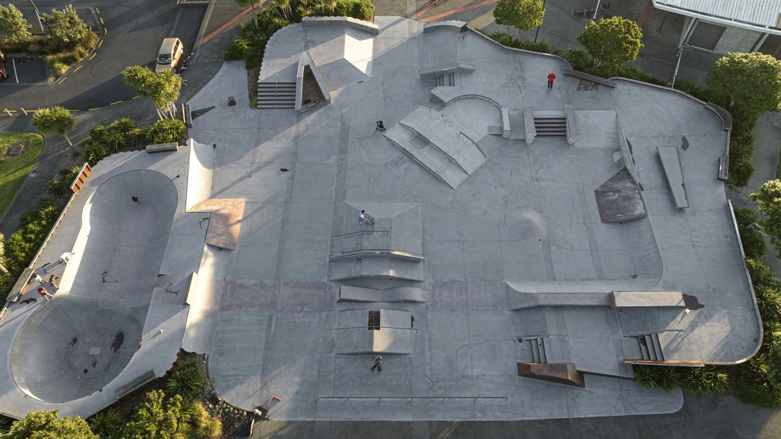 Aerial view of a concrete skatepark with multiple ramps, stairs, and rails, surrounded by trees, bushes, and parking lot, with several people skating and walking.