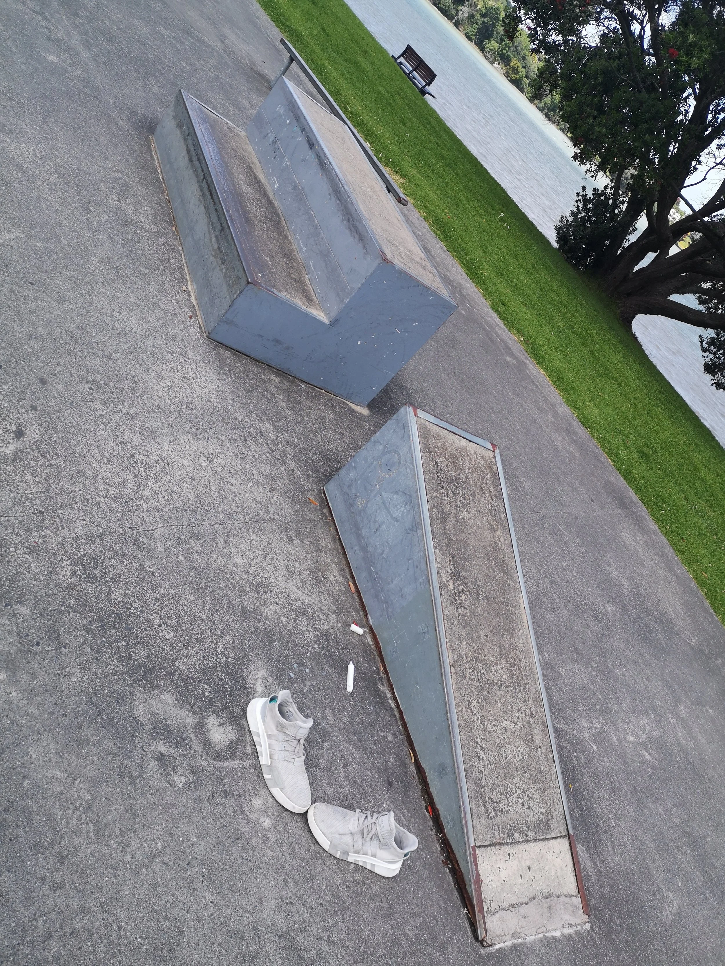 Empty skate park ledge with white sneakers on ground, near a pond with trees and bench in the background.