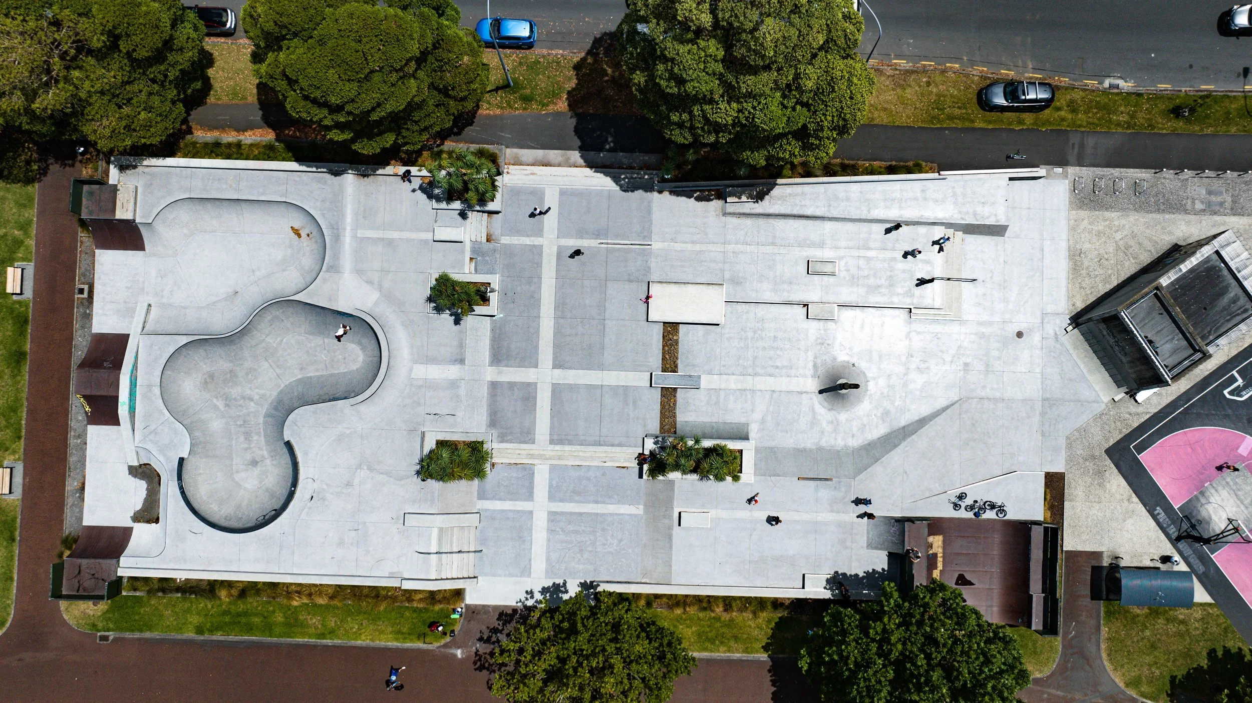 An aerial view of a skating rink and park with people walking and bikes, surrounded by trees and parked cars.