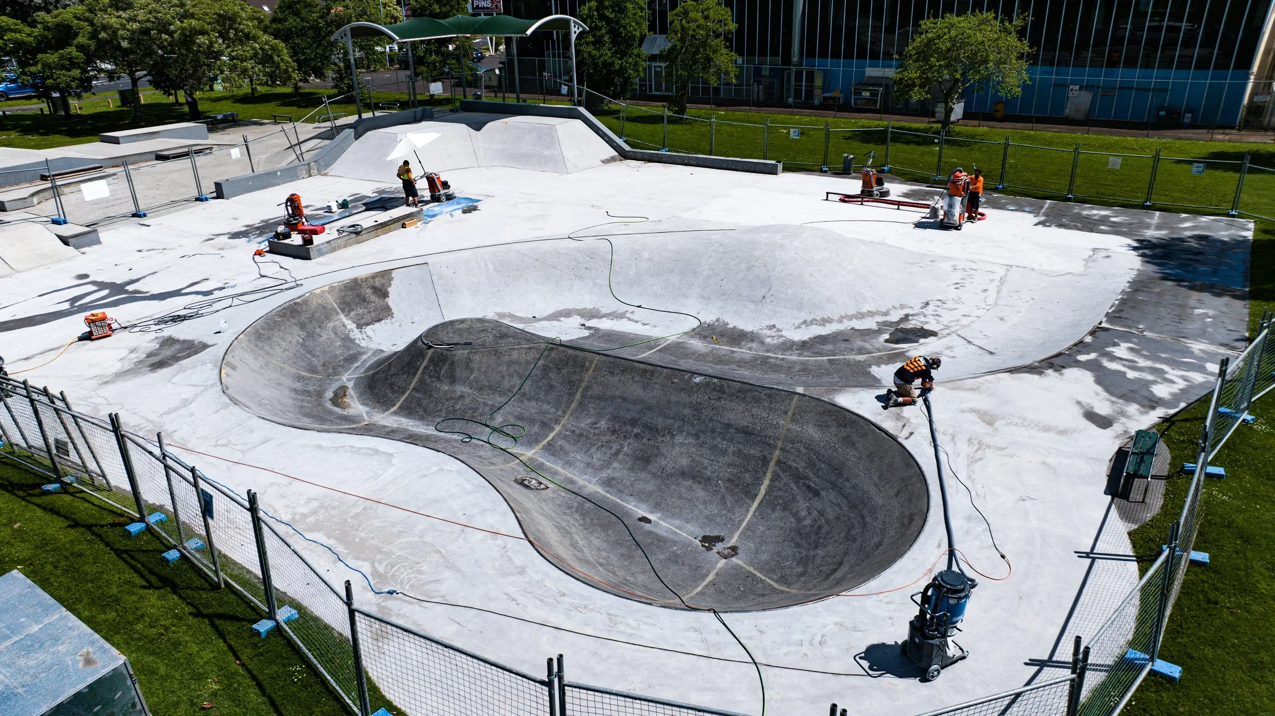Workers resurfacing a concrete skatepark with machines, surrounded by a fence and green trees, with a building and parking lot in the background.