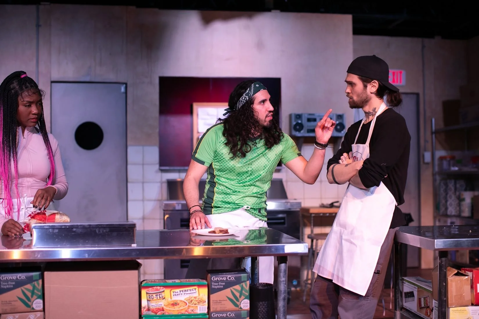 Scene in a kitchen with three people, one woman and two men, engaged in a heated discussion. The woman is on the left, wearing a pink jacket, holding a bread loaf. The man in the middle with long curly hair, a bandana, and a green shirt, is pointing his finger at the man on the right, who is wearing a black cap, black shirt, and apron, with his arms crossed. The kitchen has stainless steel counters, boxes of food, and a large TV behind them.