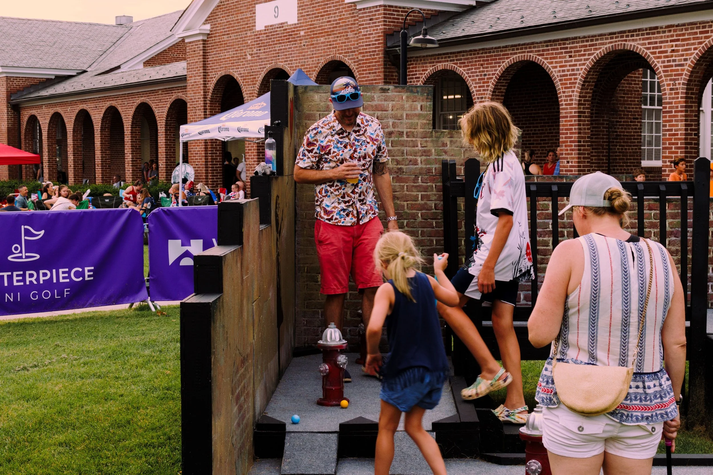 People playing mini-golf outdoors near a brick building during a summer event, with one person holding a drink and children engaging in the game.