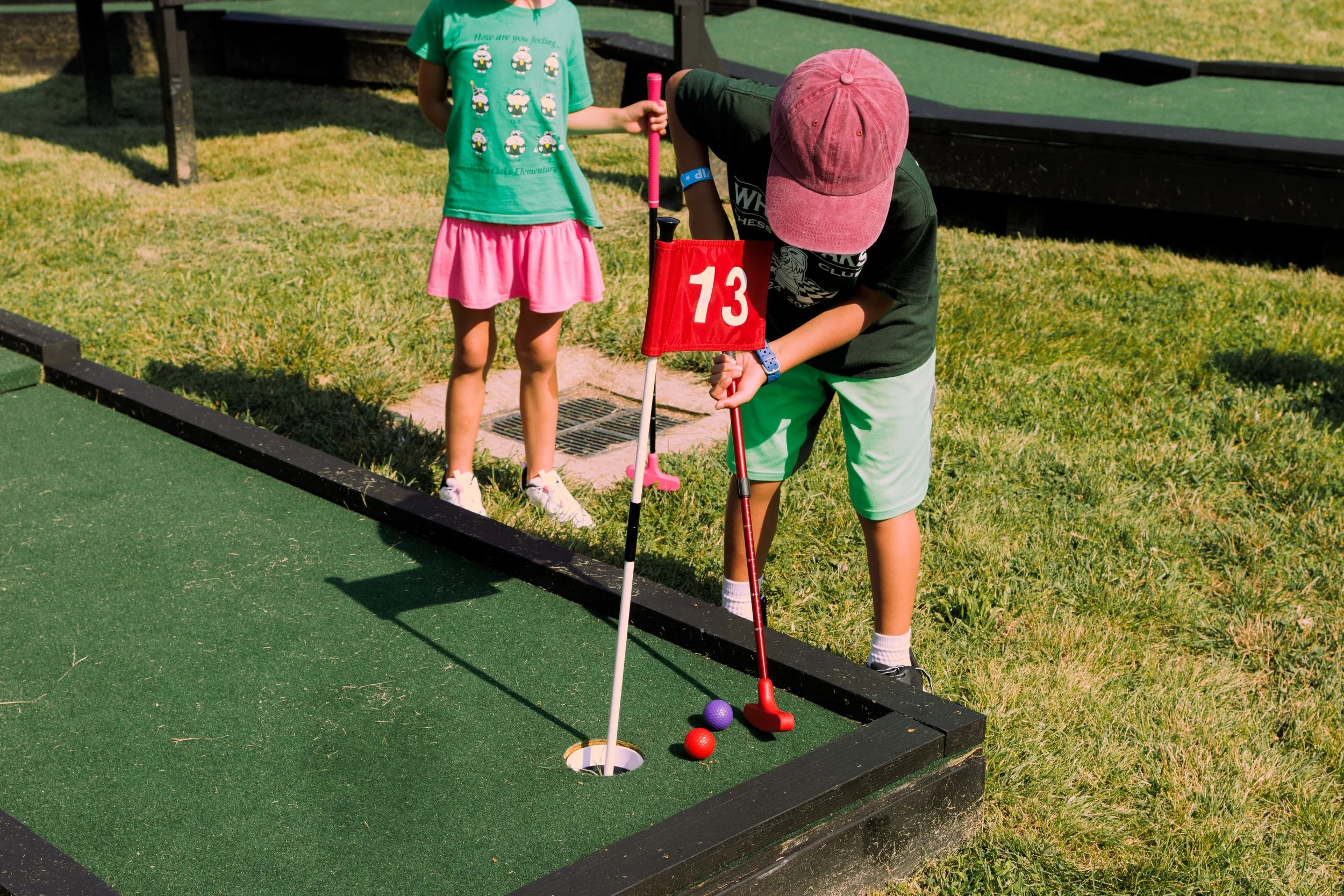 A young boy playing miniature golf, bent over aiming a red putter at a purple golf ball, near a hole marked number 13. A young girl in a green shirt and pink skirt stands nearby on the grass, observing.