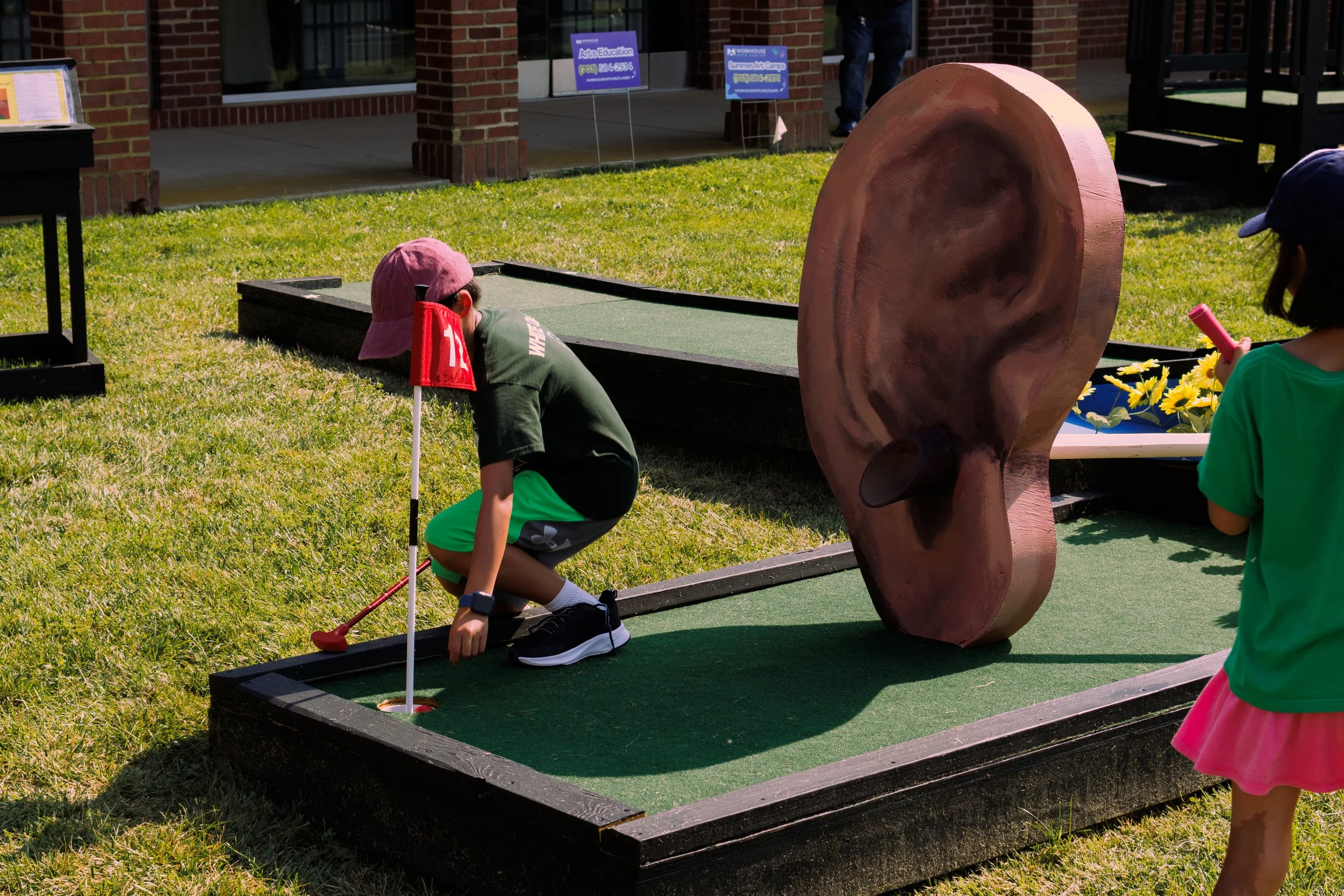 Children playing miniature golf outdoors near a large ear sculpture exhibit.