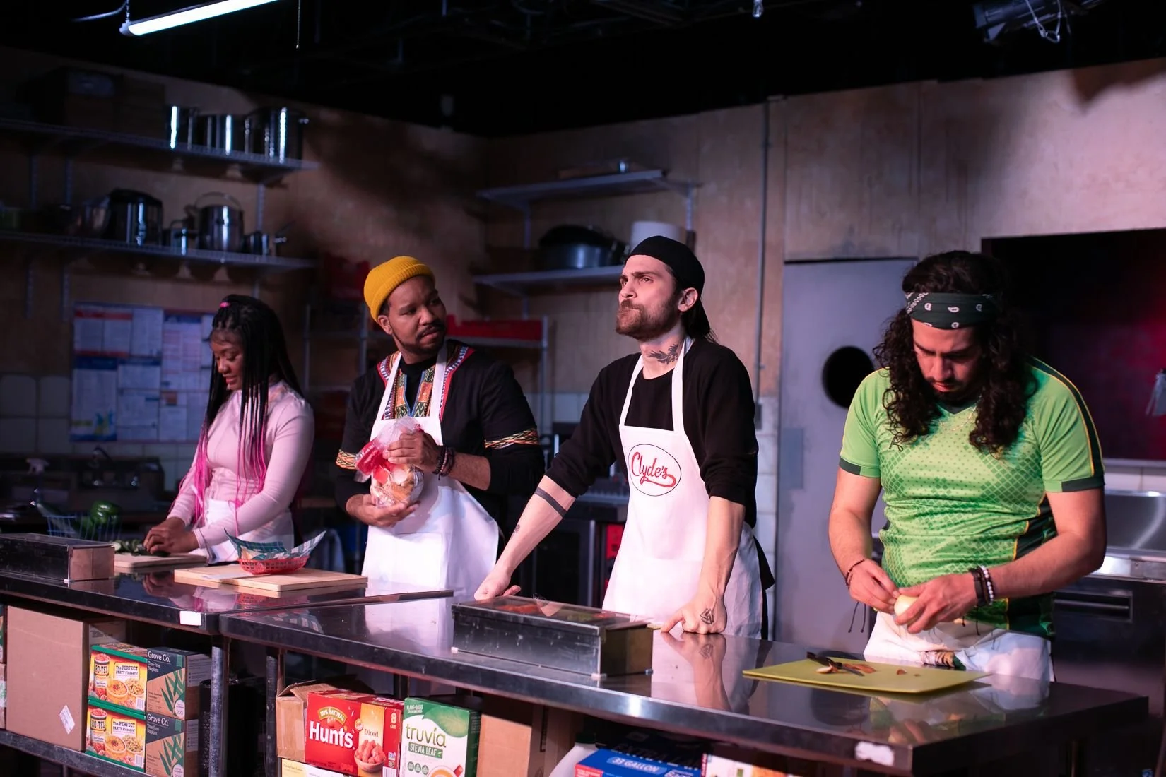 Four people in a kitchen with cooking ingredients and boxes on the counter, some wearing aprons, one chopping a vegetable, another looking contemplative, and others involved in food preparation.