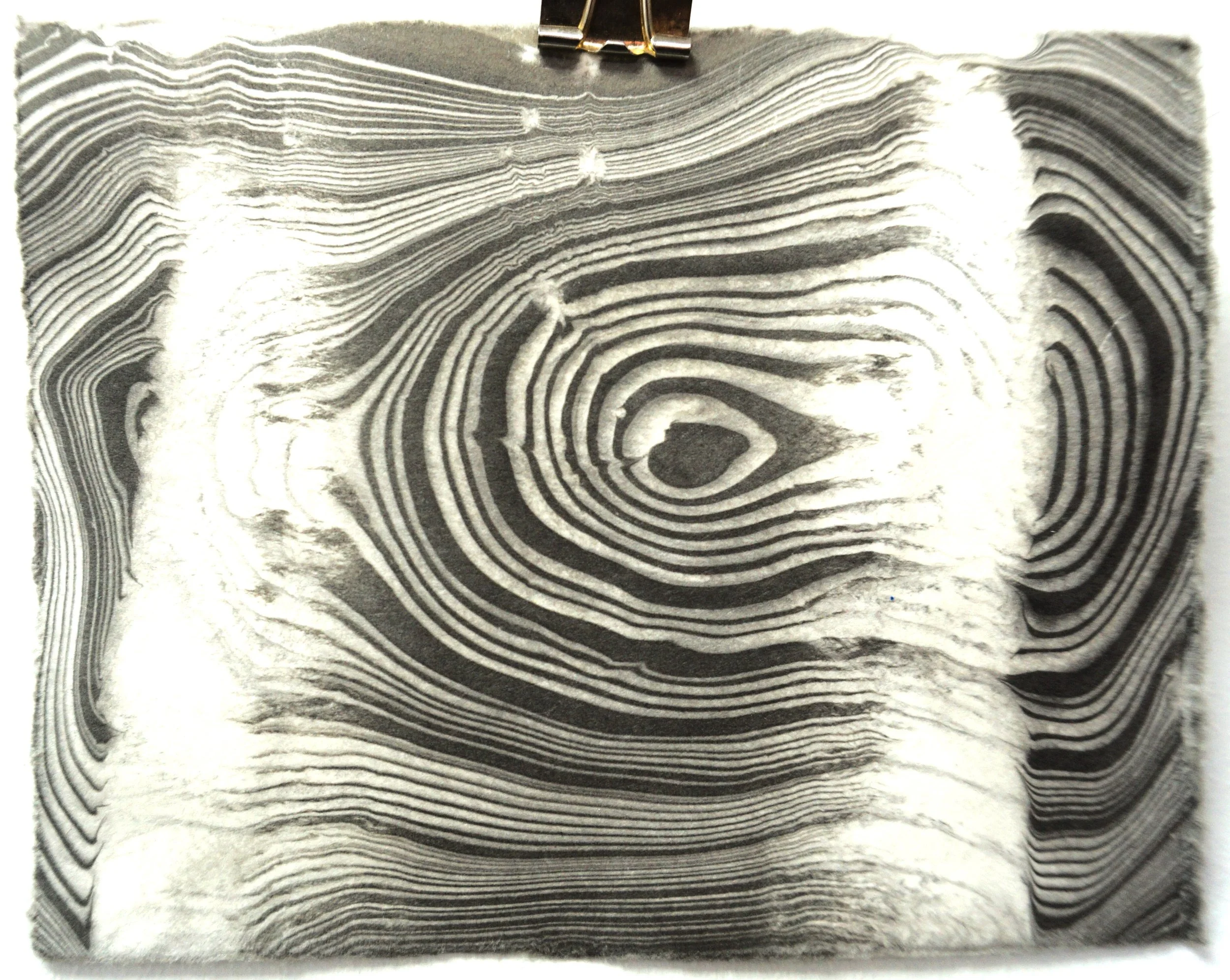A black and white close-up of a textured carved wooden surface with concentric rings and a heart-shaped pattern at the center.