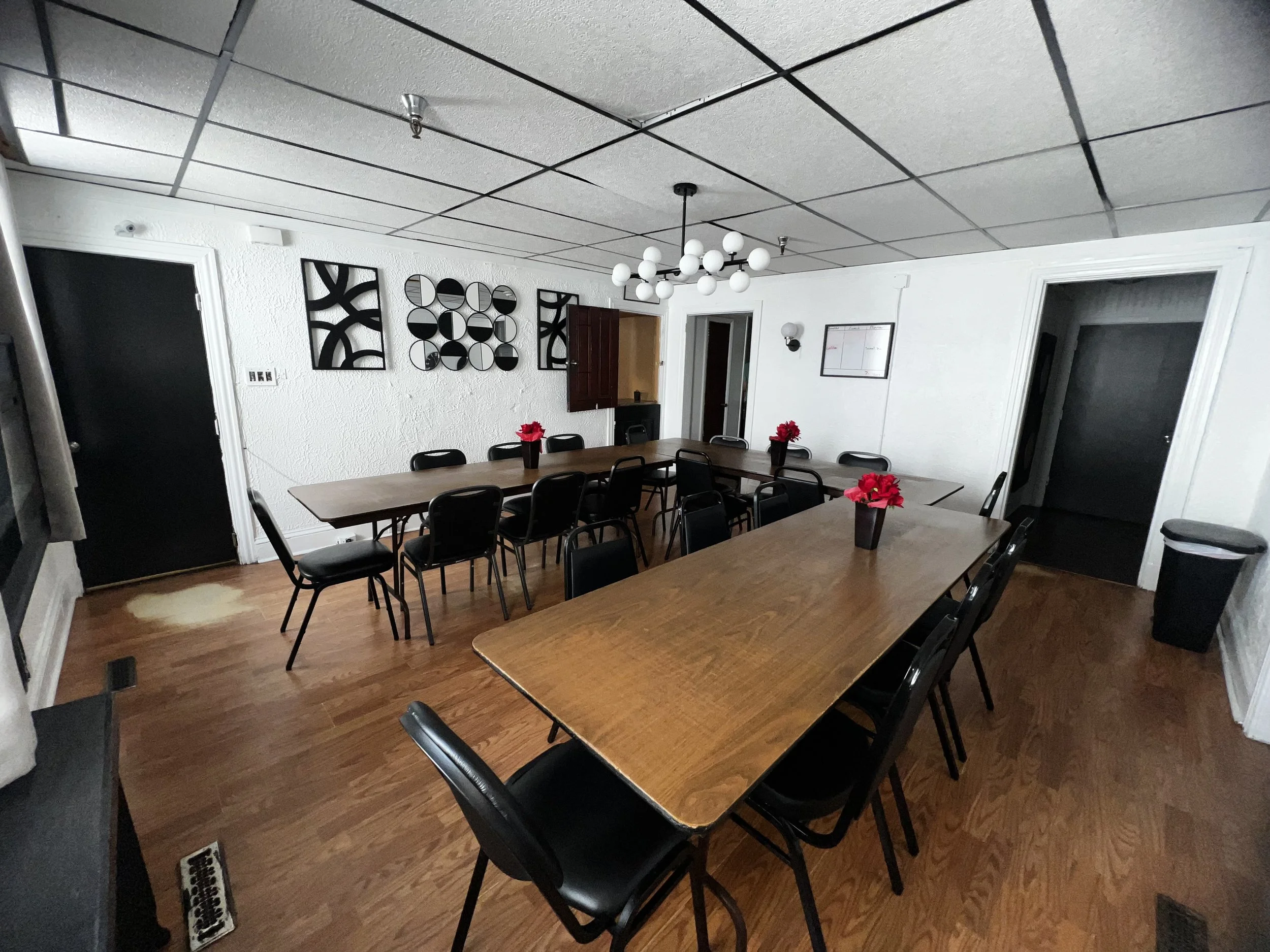 A meeting room with multiple rectangular tables surrounded by black chairs. There are red flower pots on the tables. The room has white textured walls with black geometric wall art and a whiteboard. The ceiling has large white tiles with black grid l