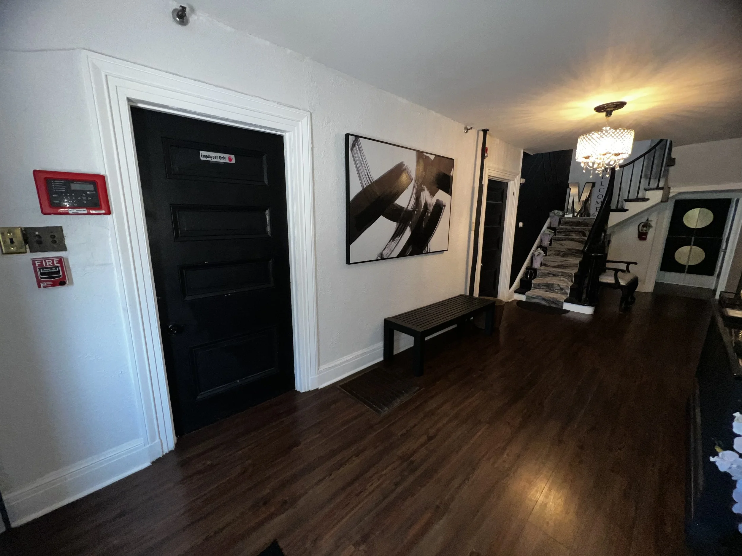 Interior of a home with a black front door labeled 'Employees Only', a painting on the wall, a staircase with patterned carpeting, a chandelier, a bench, a small black table, and a fire alarm on the wall.