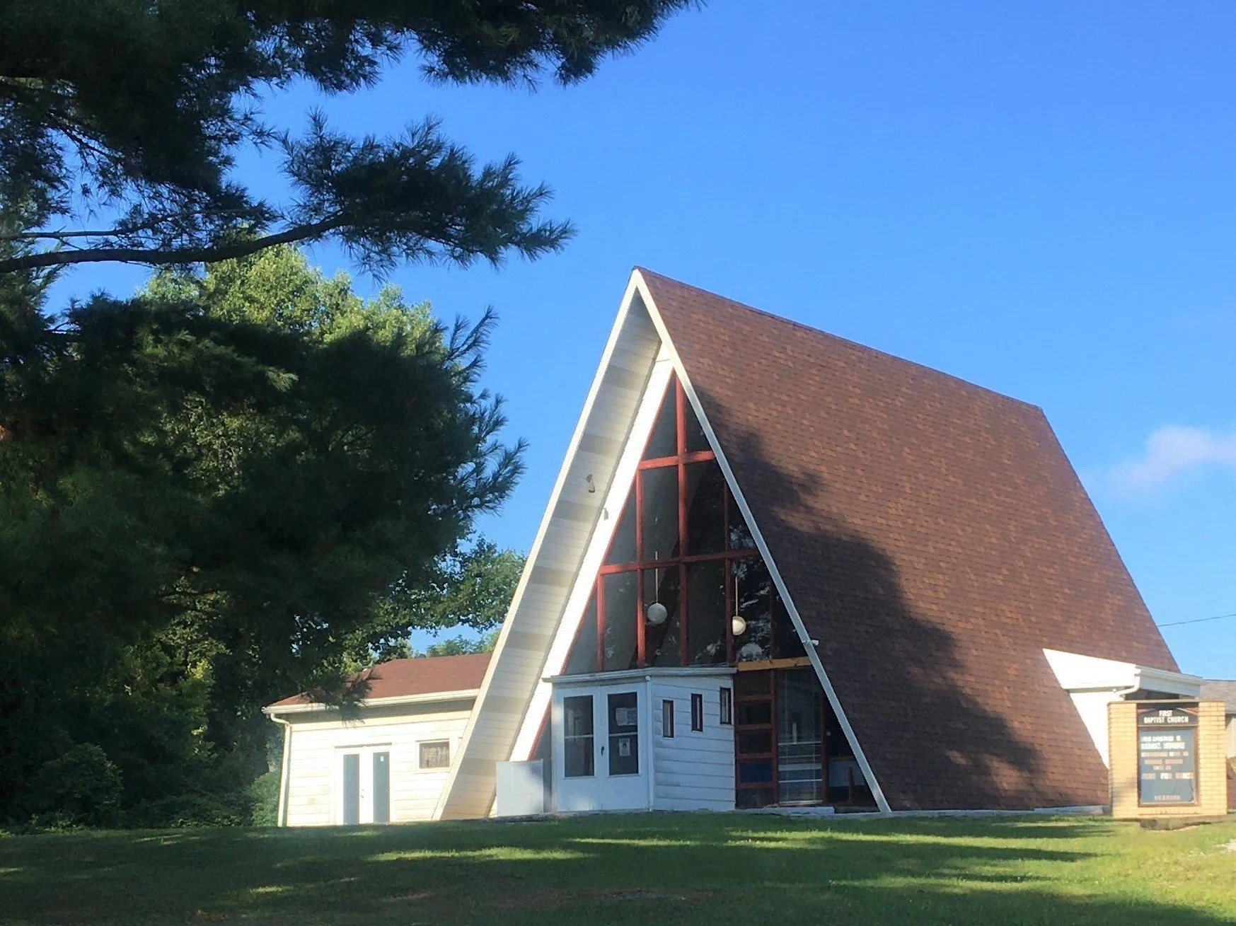 The image shows a church building with a steep, triangular roof and a large cross on the front. There are trees surrounding the church and a bright blue sky in the background.
