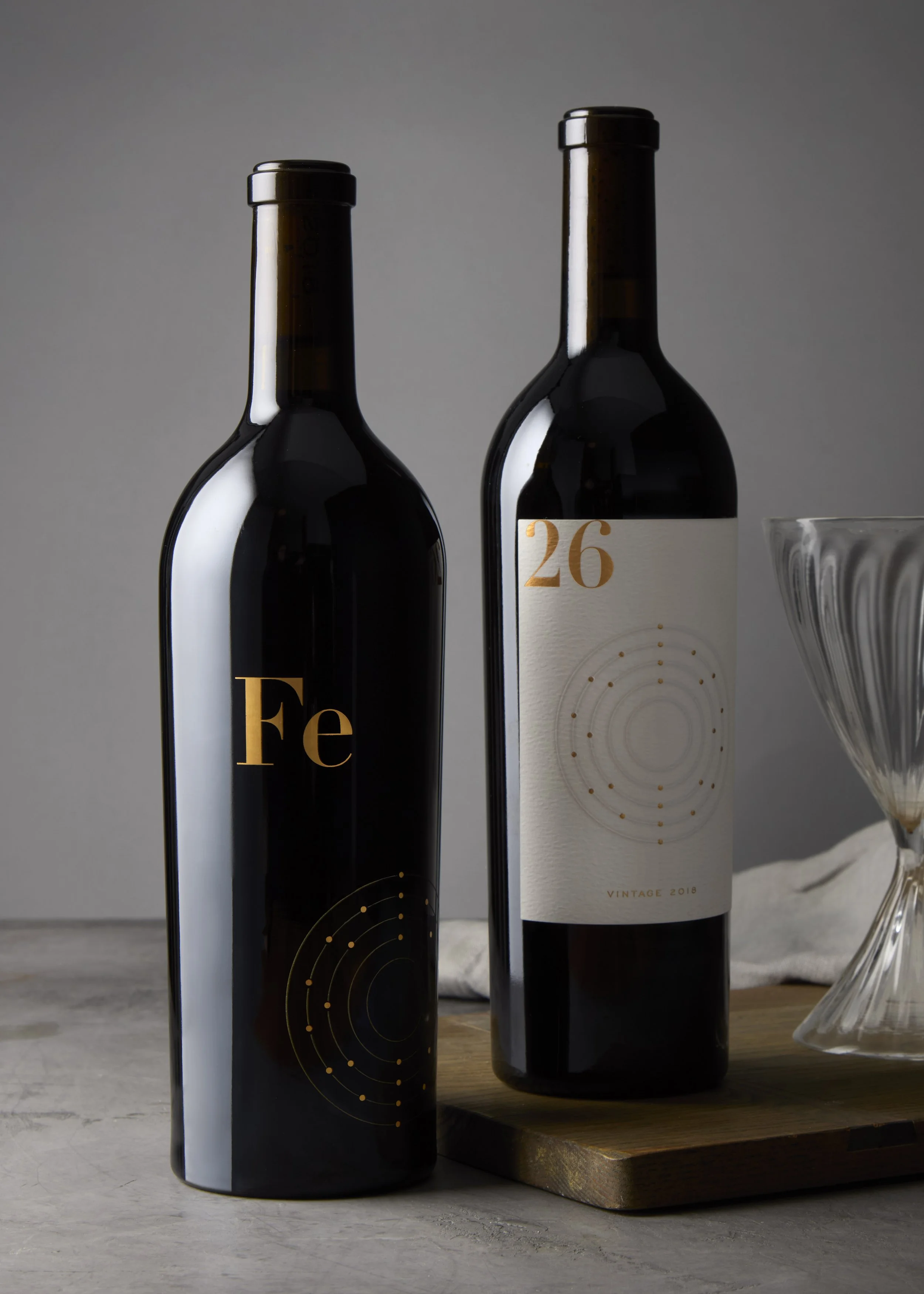 Two bottles of wine with dark glass and minimalist labels, one white and one red, placed on a light-colored table near a wooden cutting board and a vintage wine glass.