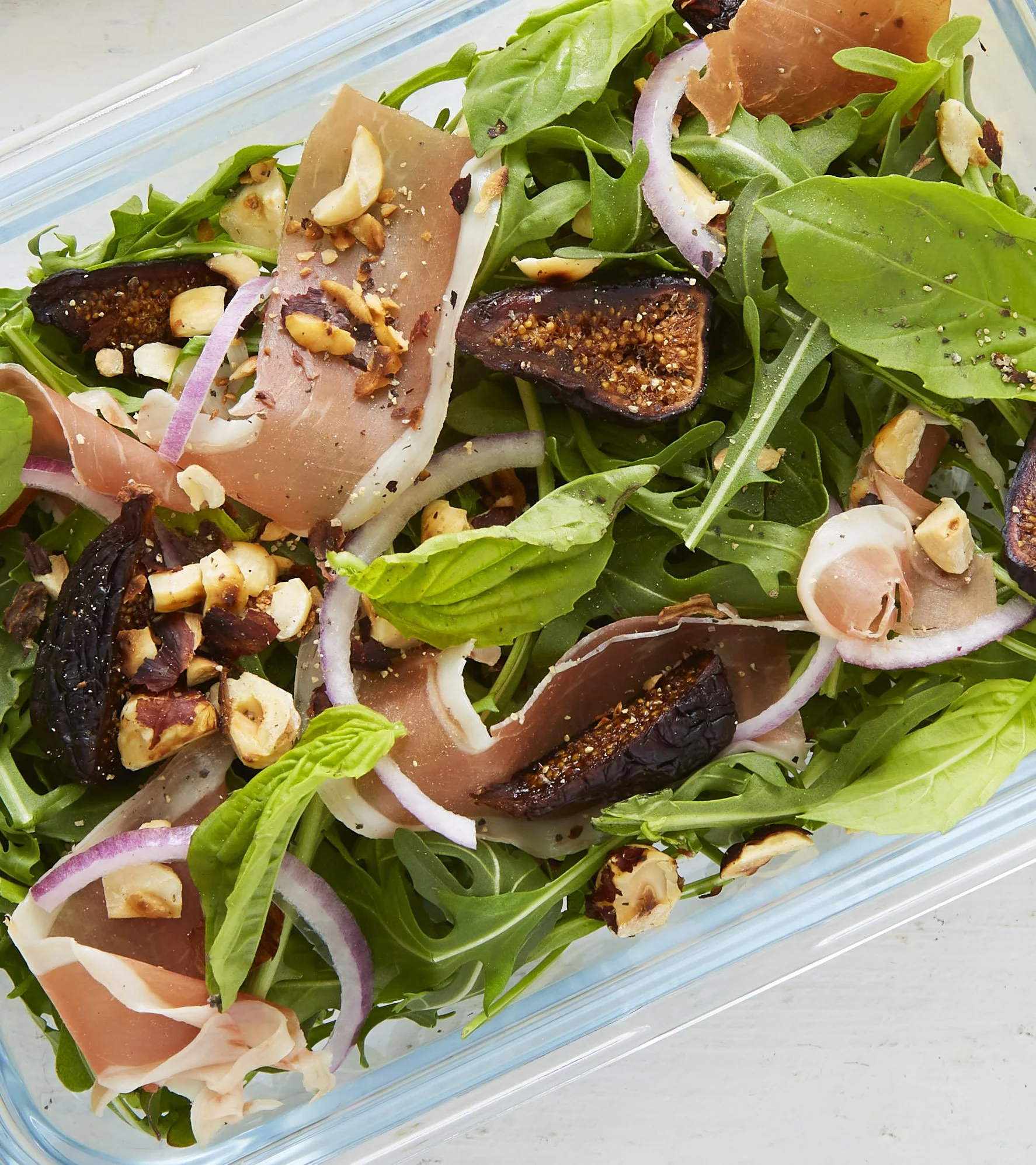 Fresh arugula salad with sliced red onions, dried figs, prosciutto, chopped nuts, and cracked black pepper in a glass container.