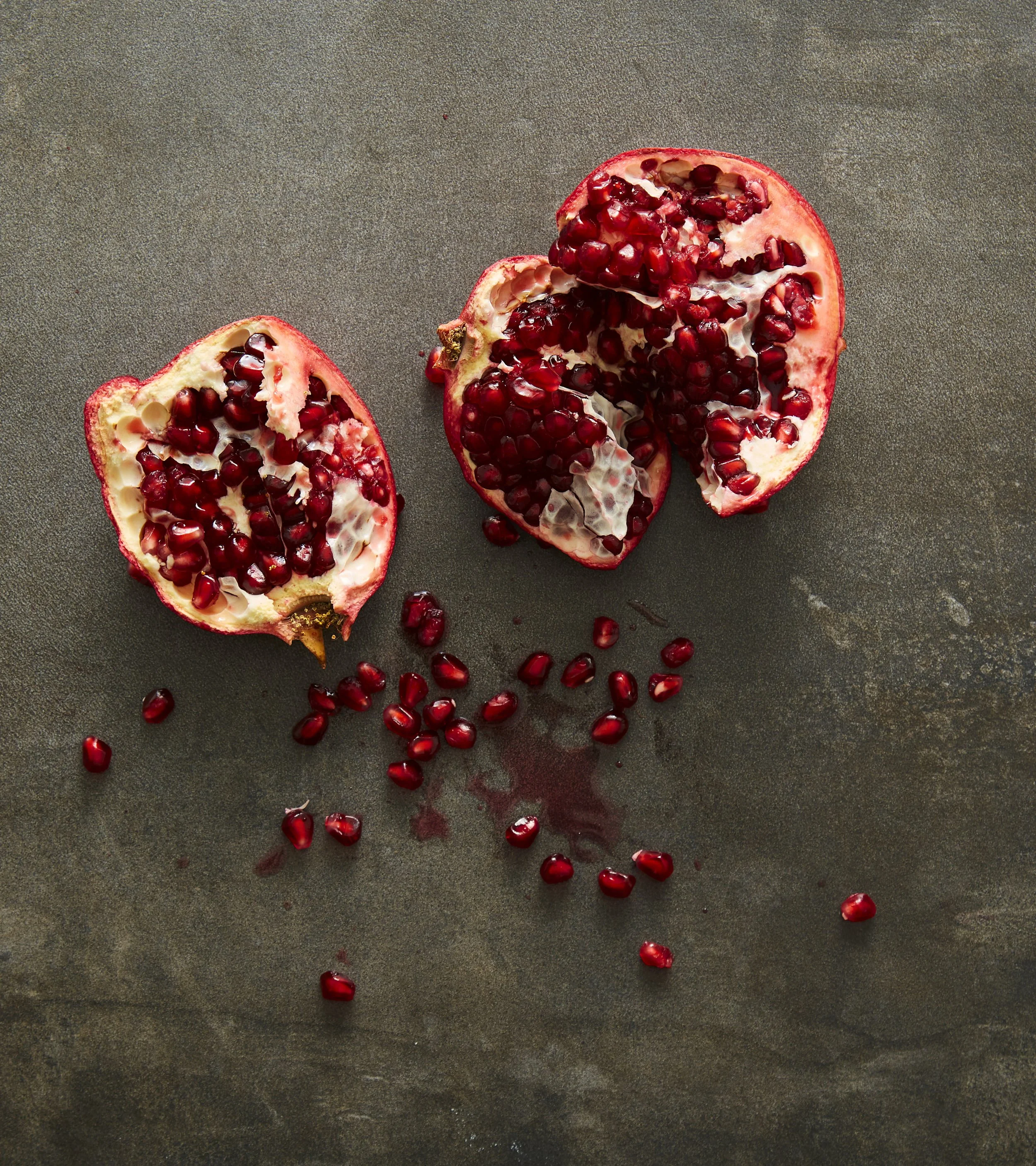 Quartered pomegranate with red seeds scattered on a dark textured surface.