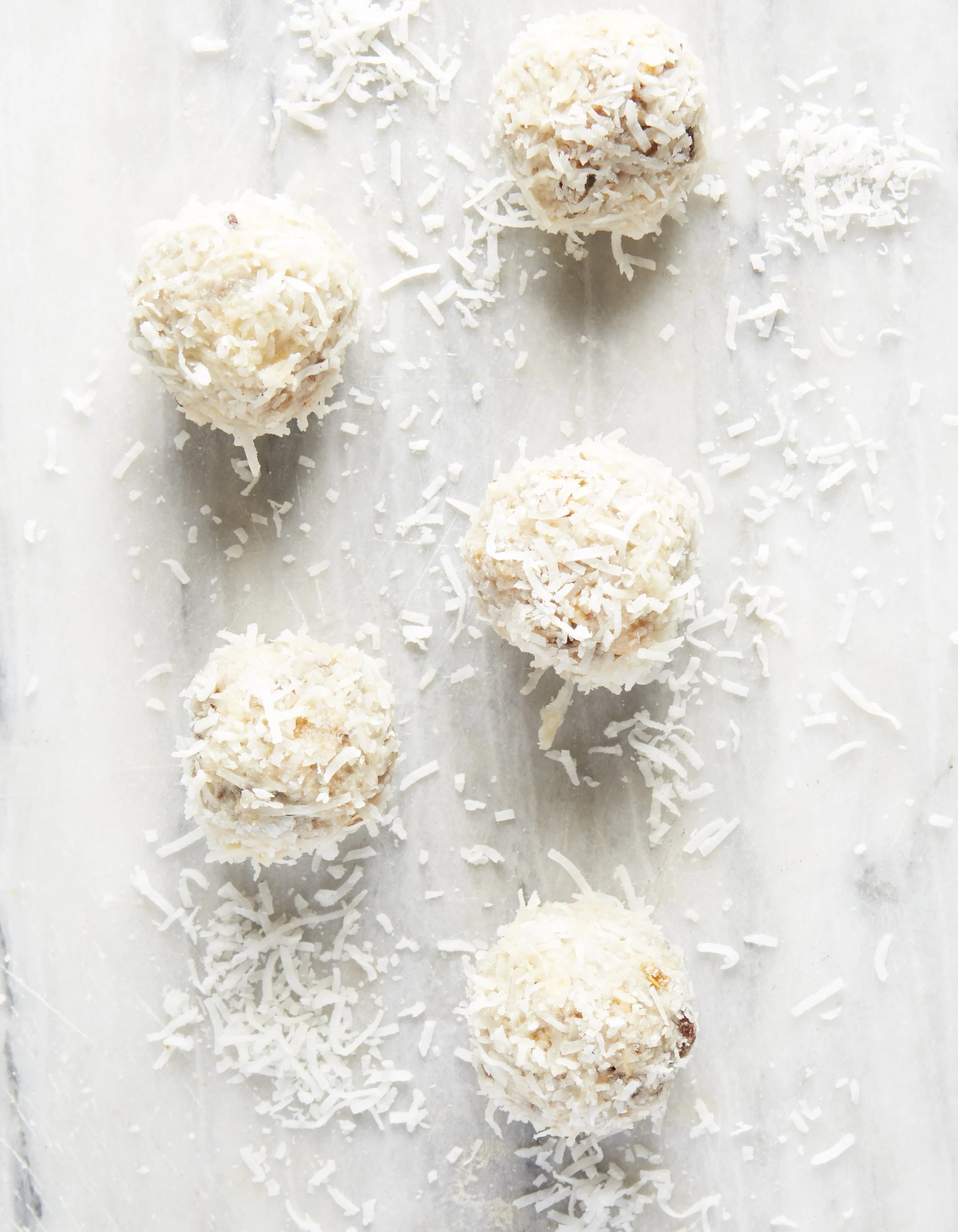 Six round white coconut-covered confections on a white surface, with shredded coconut scattered around.