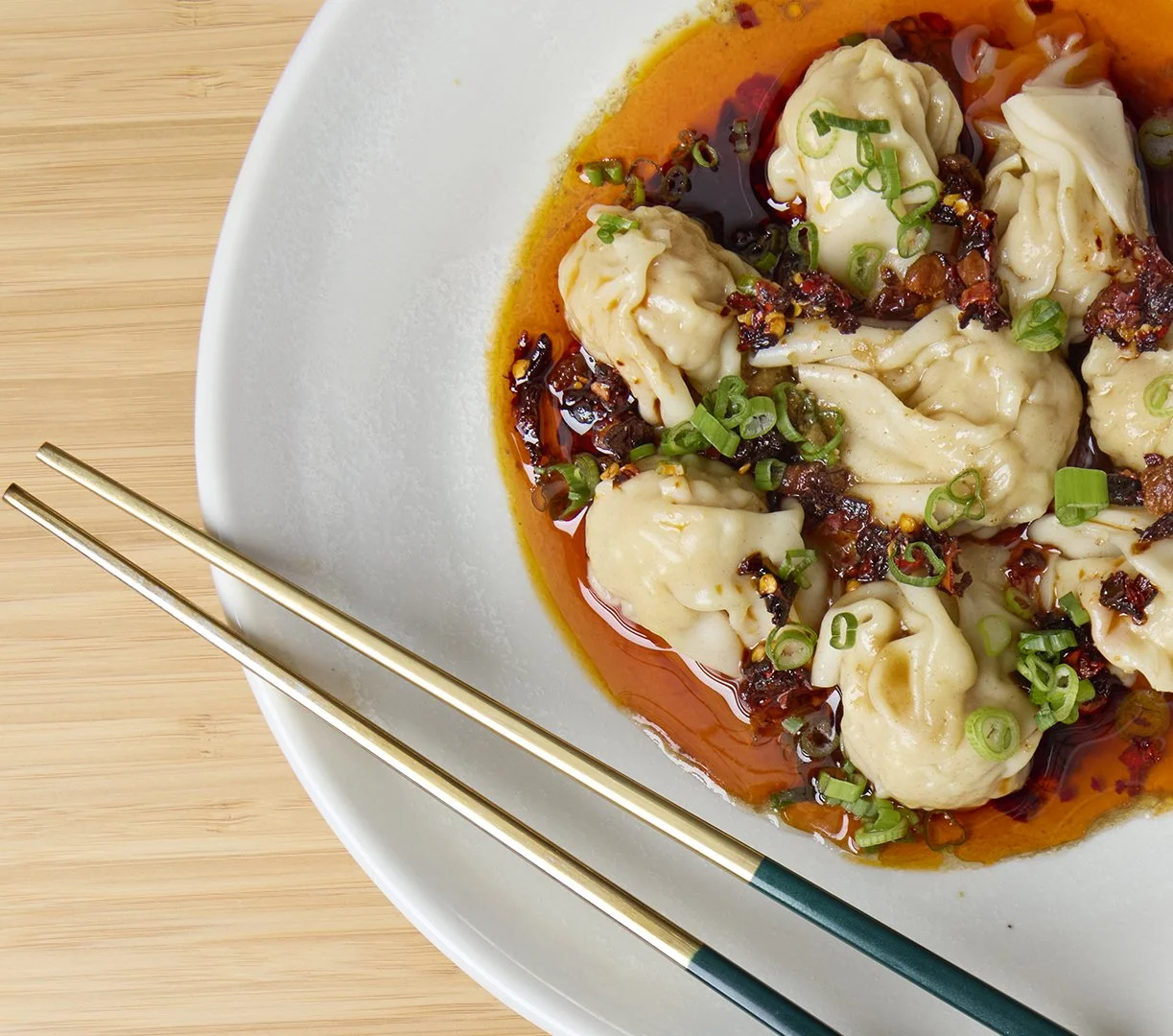 Nicole Gomes Canada's Allstar Top Chef new restaurant Lucky Lily Asian dumplings garnished with chopped green onions, served in a spicy red sauce on a white plate with chopsticks.
