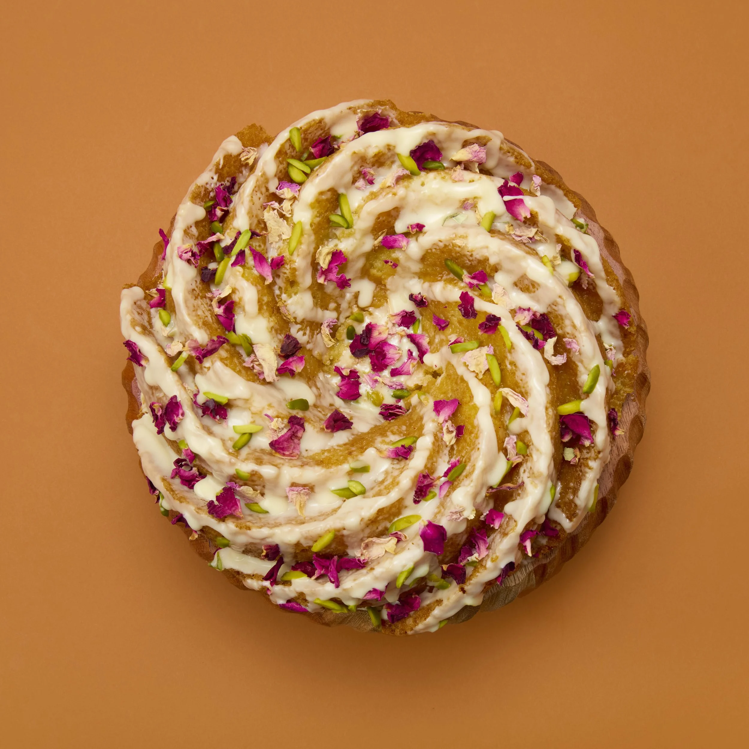 Close-up of a cinnamon roll topped with white icing, pink rose petals, and chopped green pistachios on a peach-colored background.