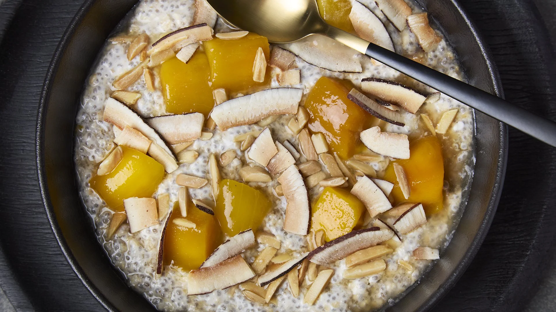 Bowl of tapioca pudding topped with sliced mango's, shredded toasted coconut, and chopped almonds, with a spoon resting inside.