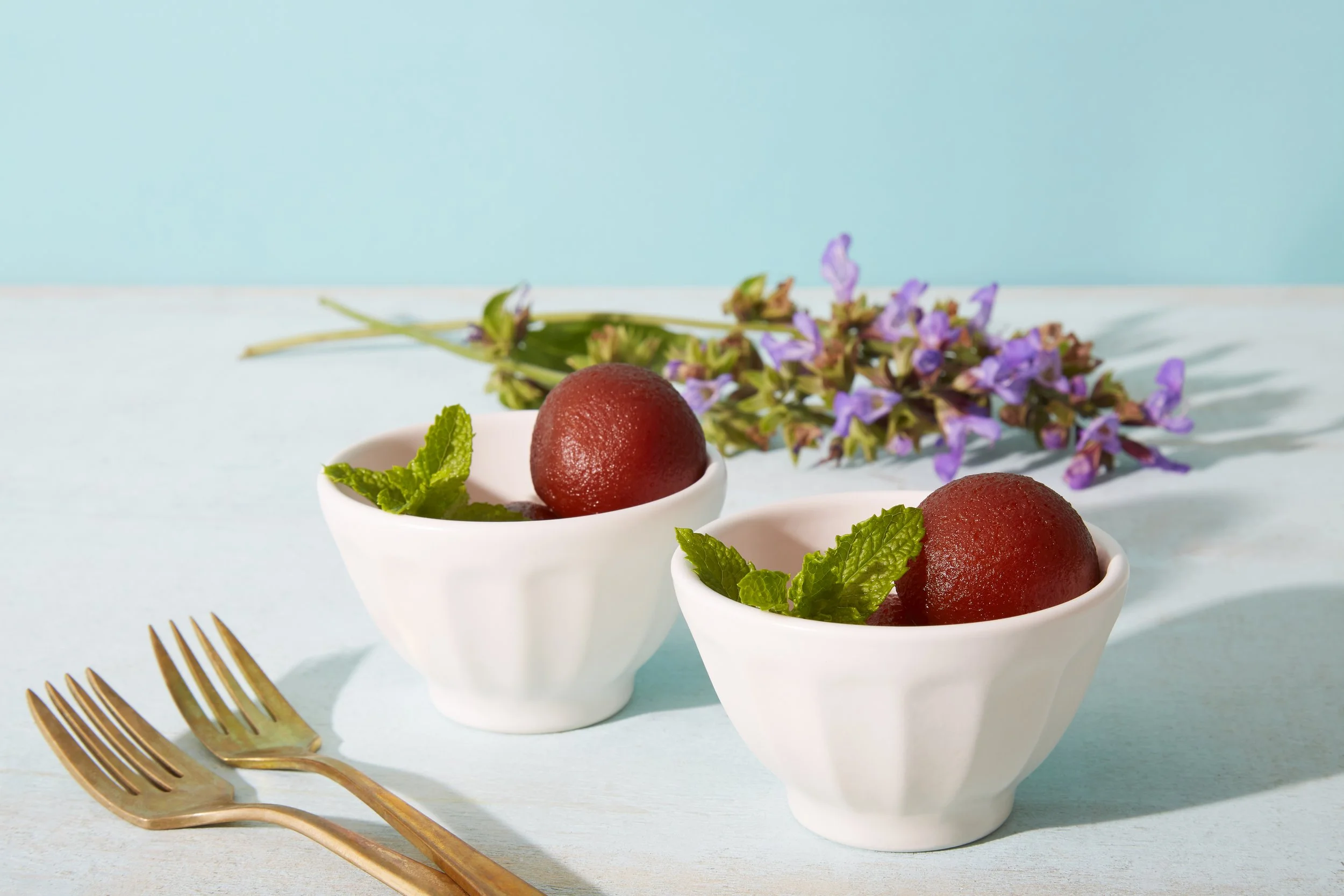 Two white bowls containing red desserts garnished with mint leaves, with a bunch of purple flowers and gold forks placed on a light blue surface against a matching light blue background.