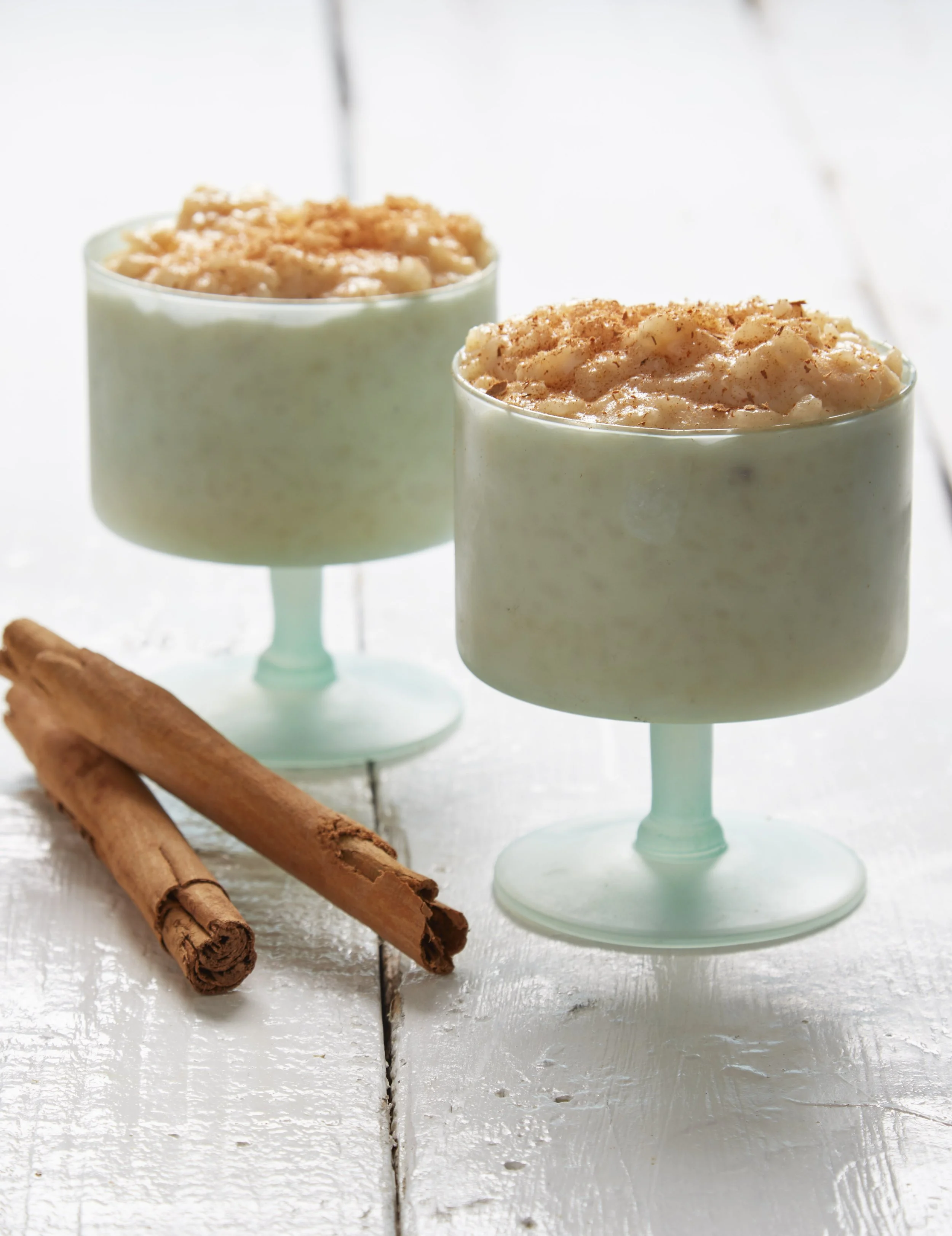 Two mint green glasses with a creamy dessert topped with crushed cookies or graham crackers, placed on a white wooden surface with cinnamon sticks nearby.