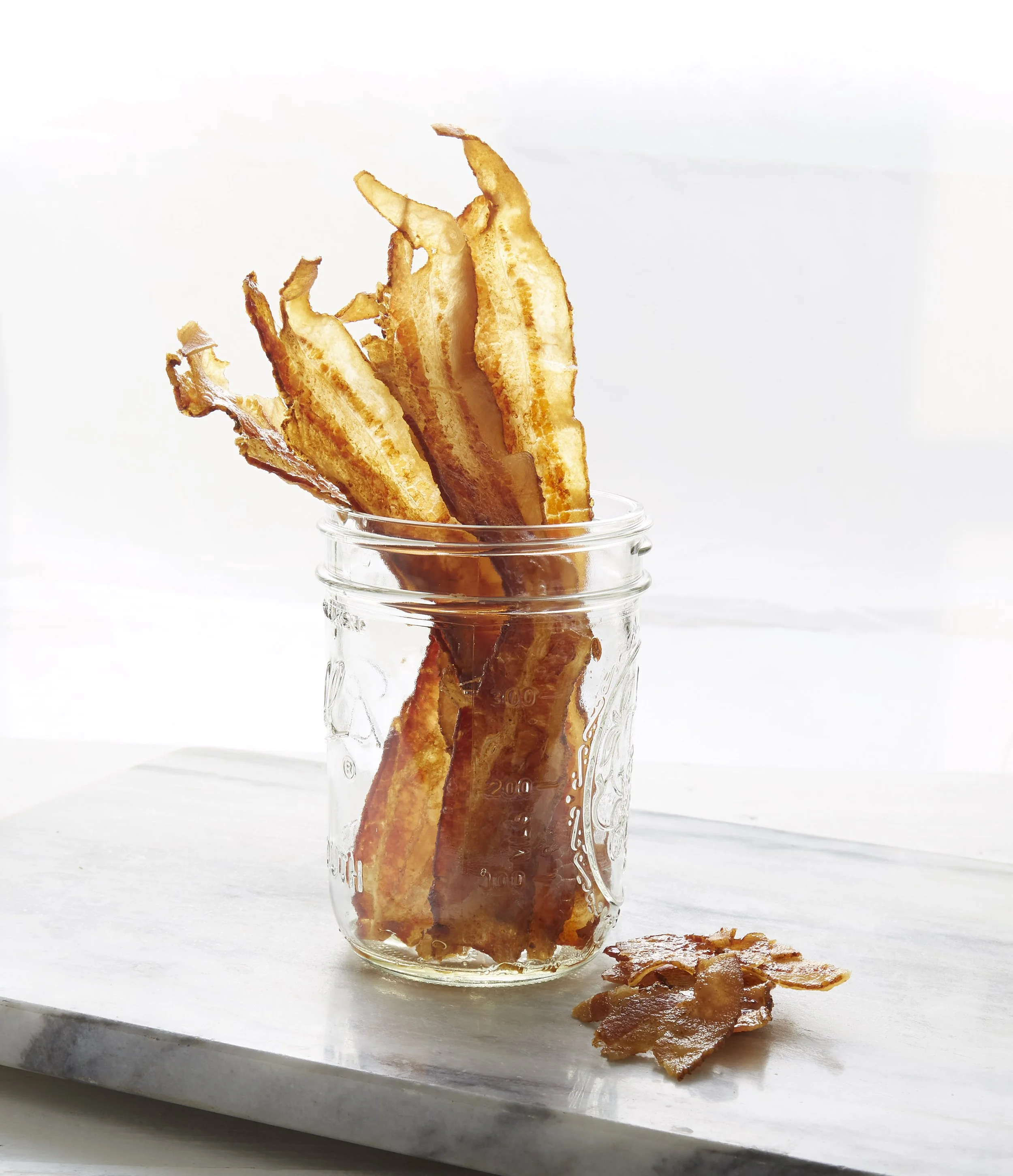 Crispy bacon strips inside a glass jar with a few bacon pieces on the white surface outside the jar.