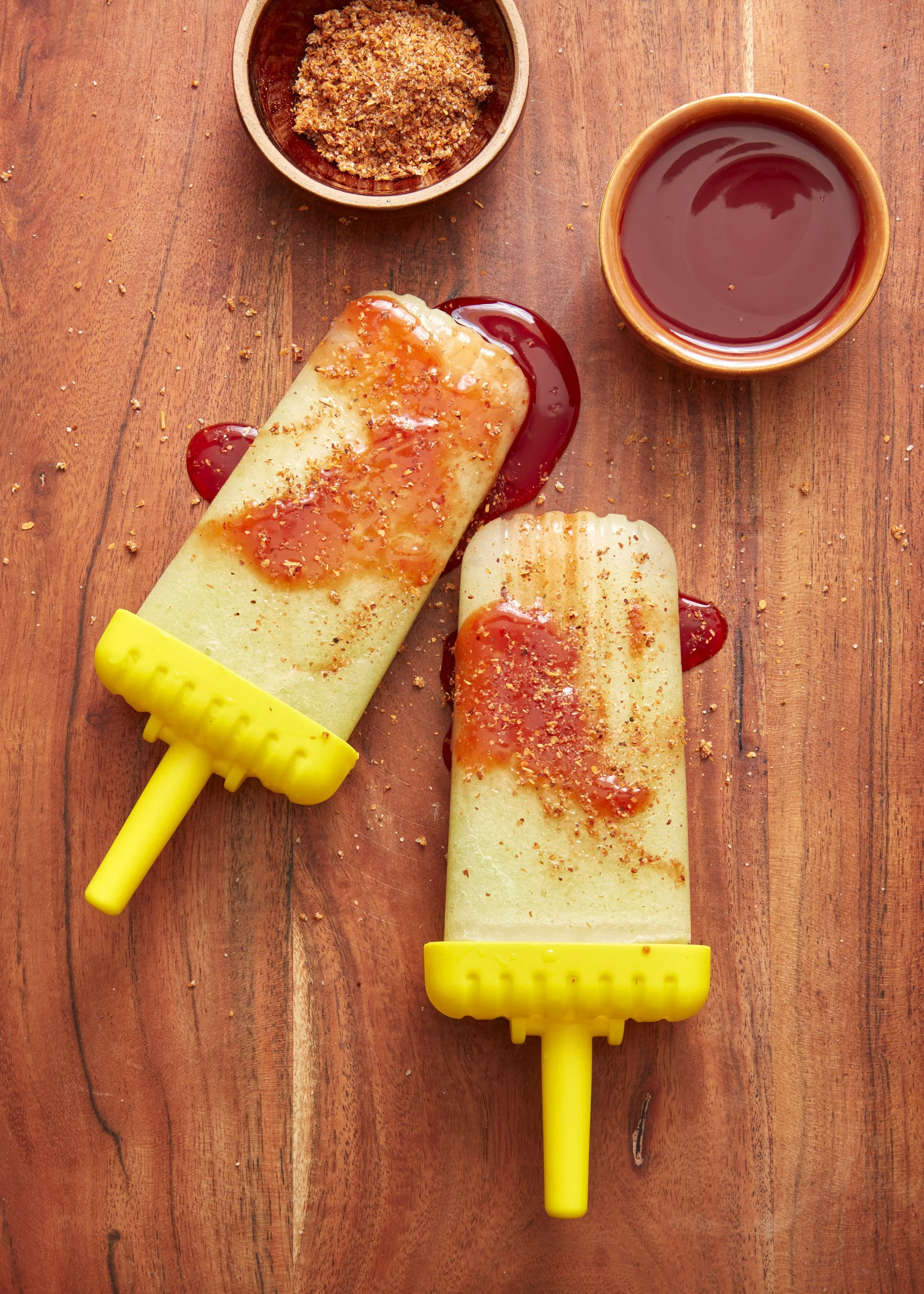 A wooden table with two frozen popsicles with yellow handles, drizzled with red syrup and sprinkled with crushed nuts, and a small bowl of crushed nuts, and a cup of dark red juice.