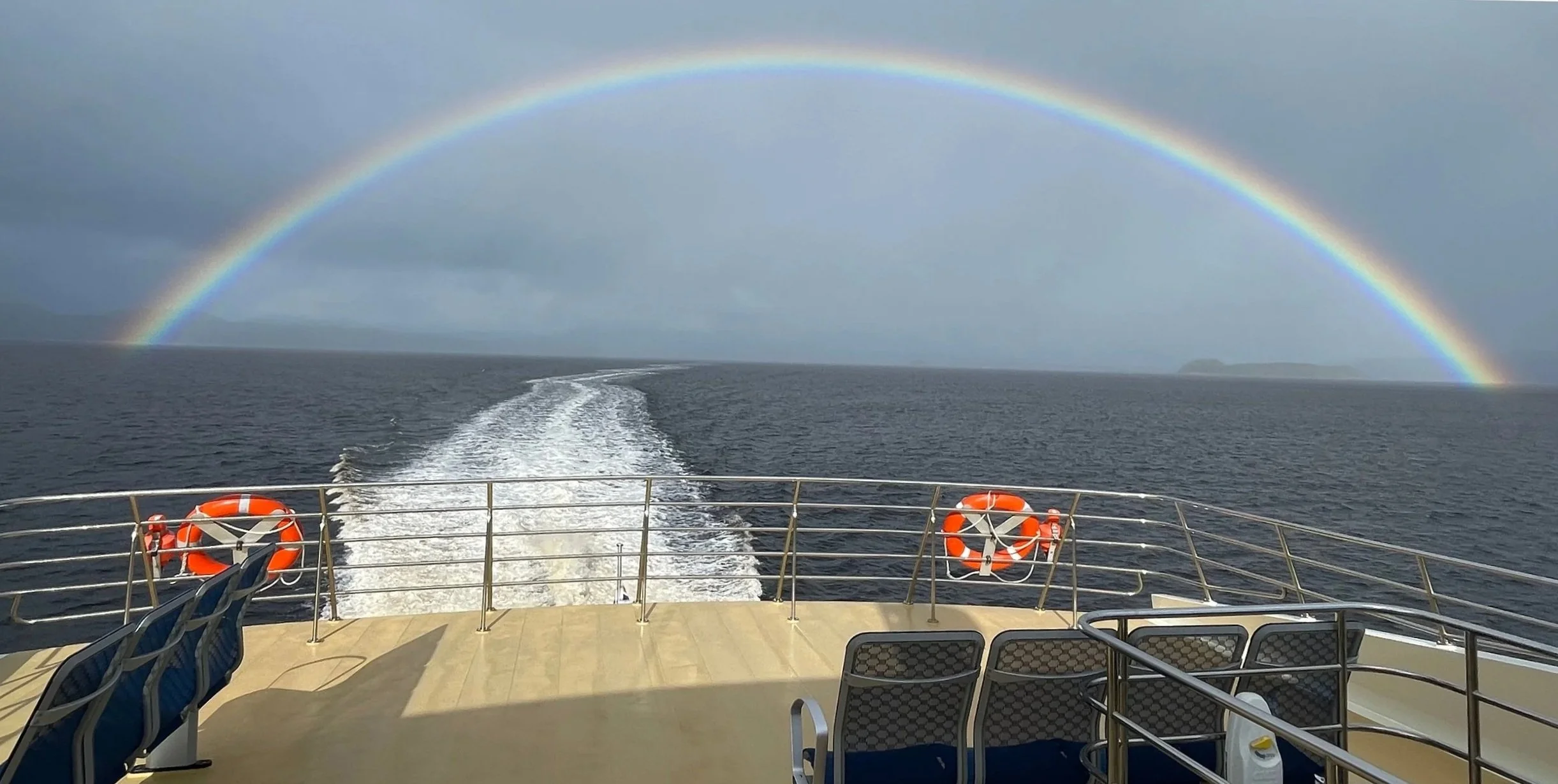View looking backward from a boat travelling on dark waters. A dark sky and rainbow above.