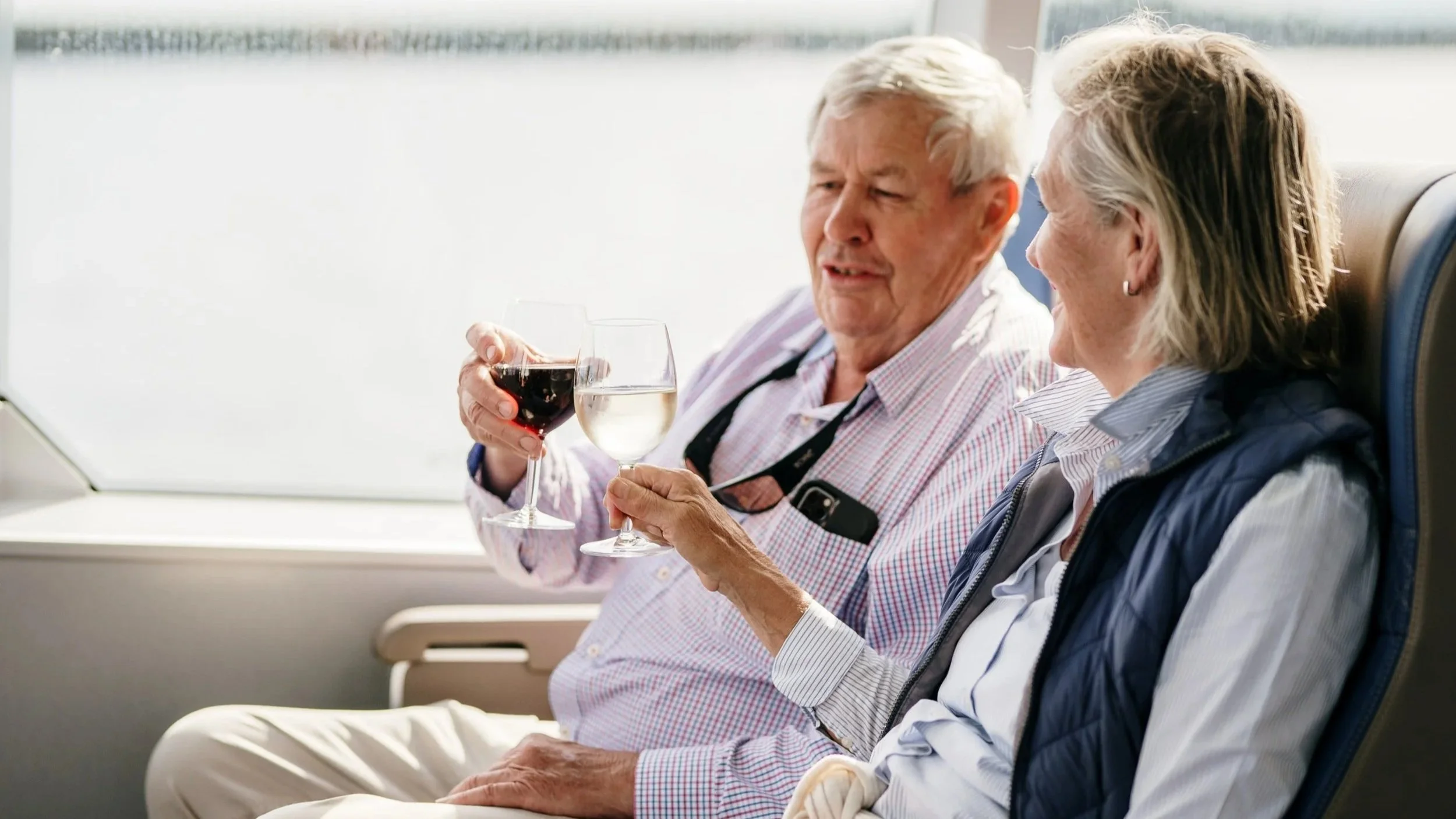 Elderly couple sitting on a boat beside a window toasting glasses of wine