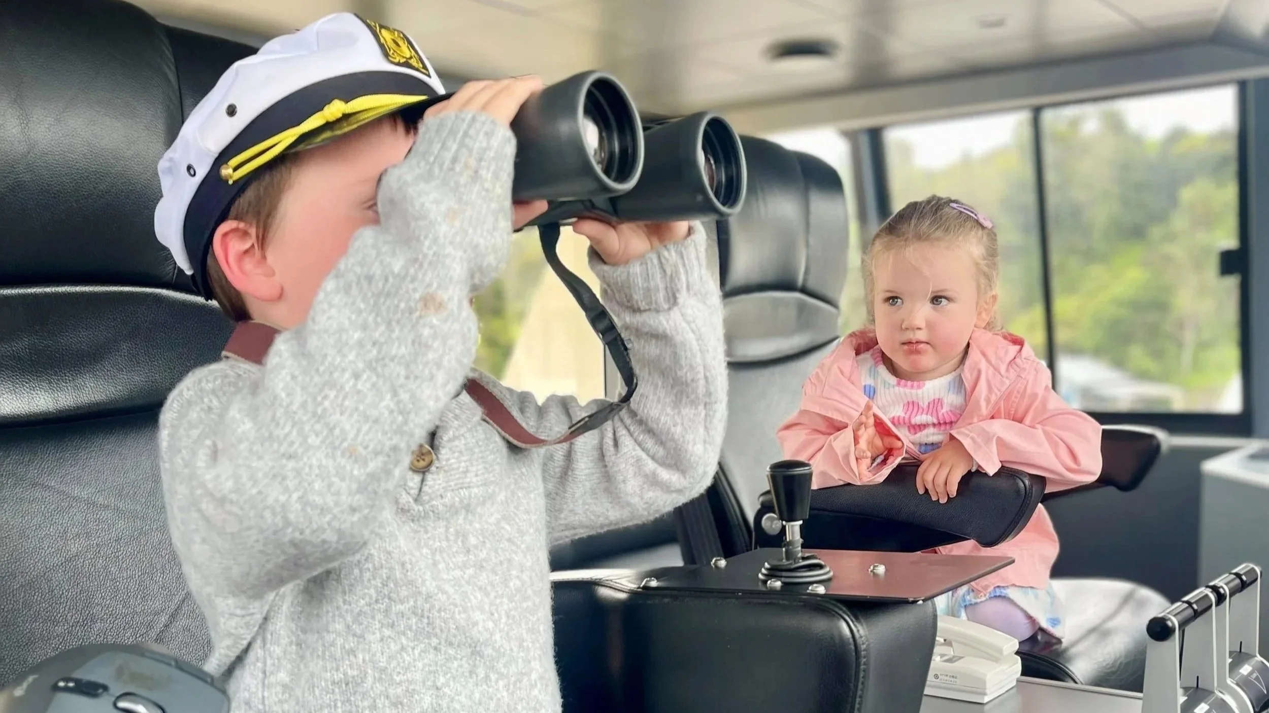 Children in the wheelhouse of a boat. One is wearing a captains hat and looking through binoculars.