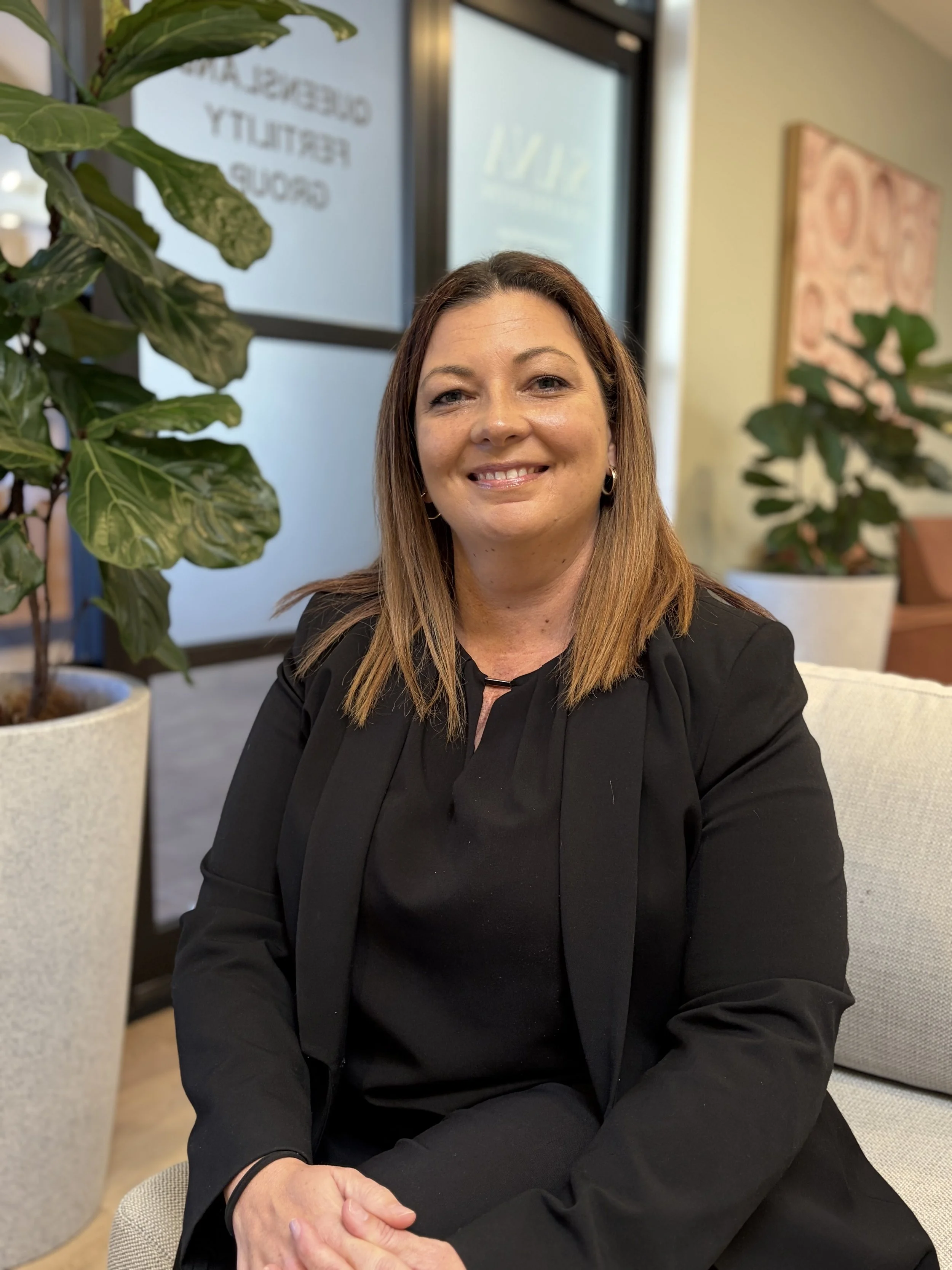 A woman with shoulder-length brown hair, wearing a black blazer, sitting on a beige sofa in an indoor setting with potted plants and a wall art, smiling at the camera.