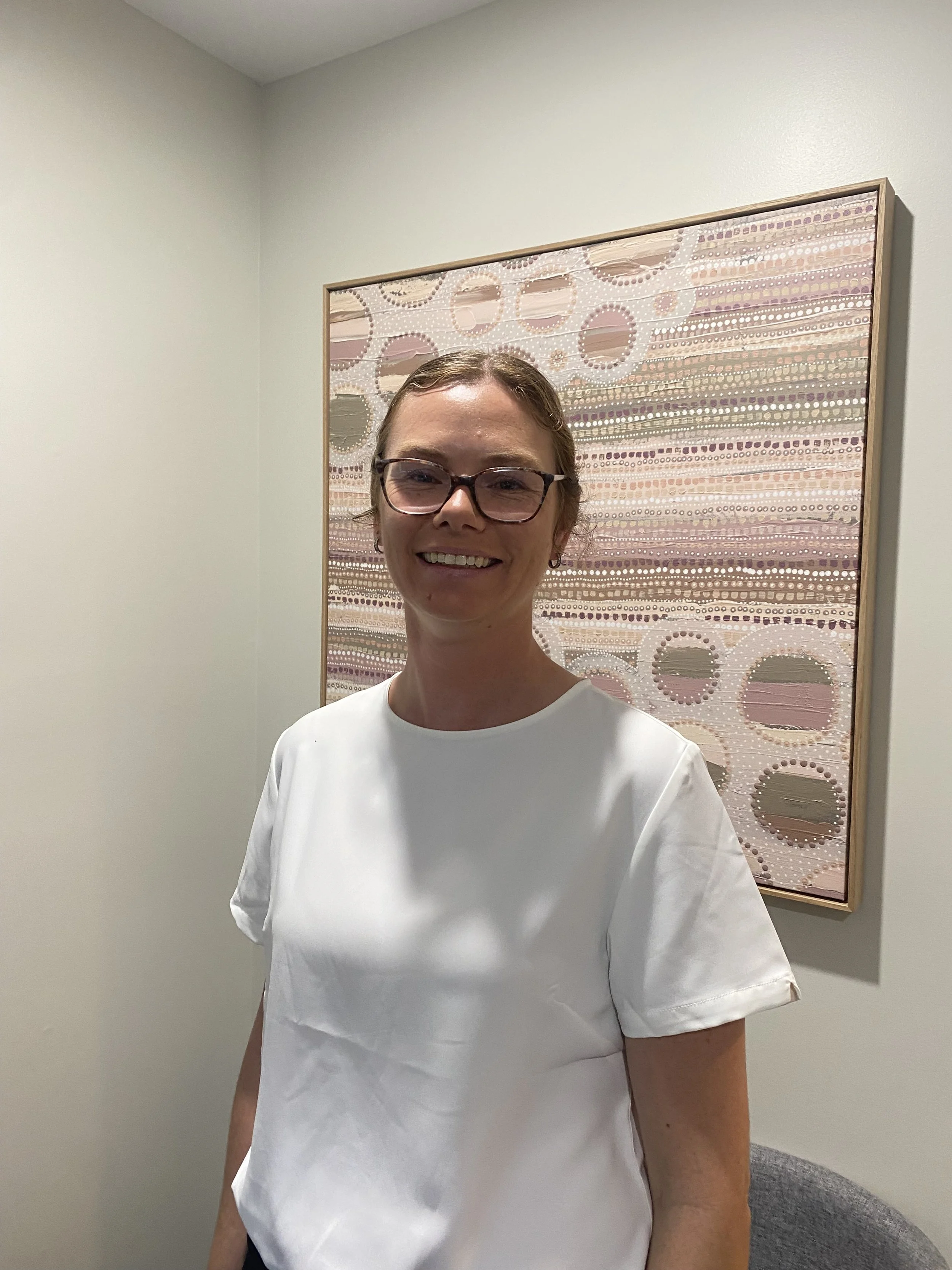A woman with glasses smiling, standing in front of an abstract art piece with circular patterns, white walls, and a gray chair