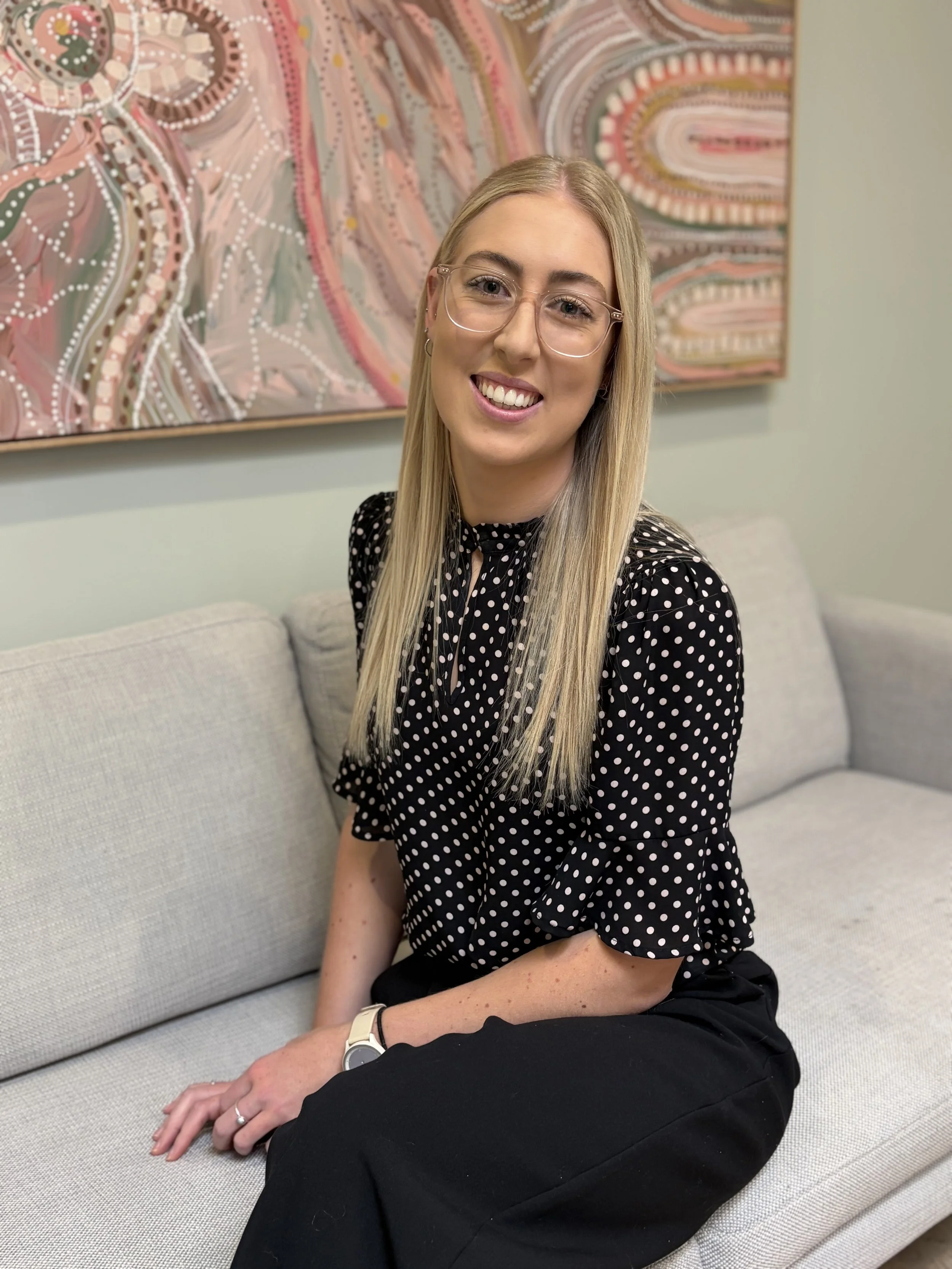A young woman with long blonde hair, wearing glasses, a black polka dot blouse, black pants, and a white wristwatch, is sitting on a light-colored sofa with a colorful abstract painting in pink, beige, and white behind her.