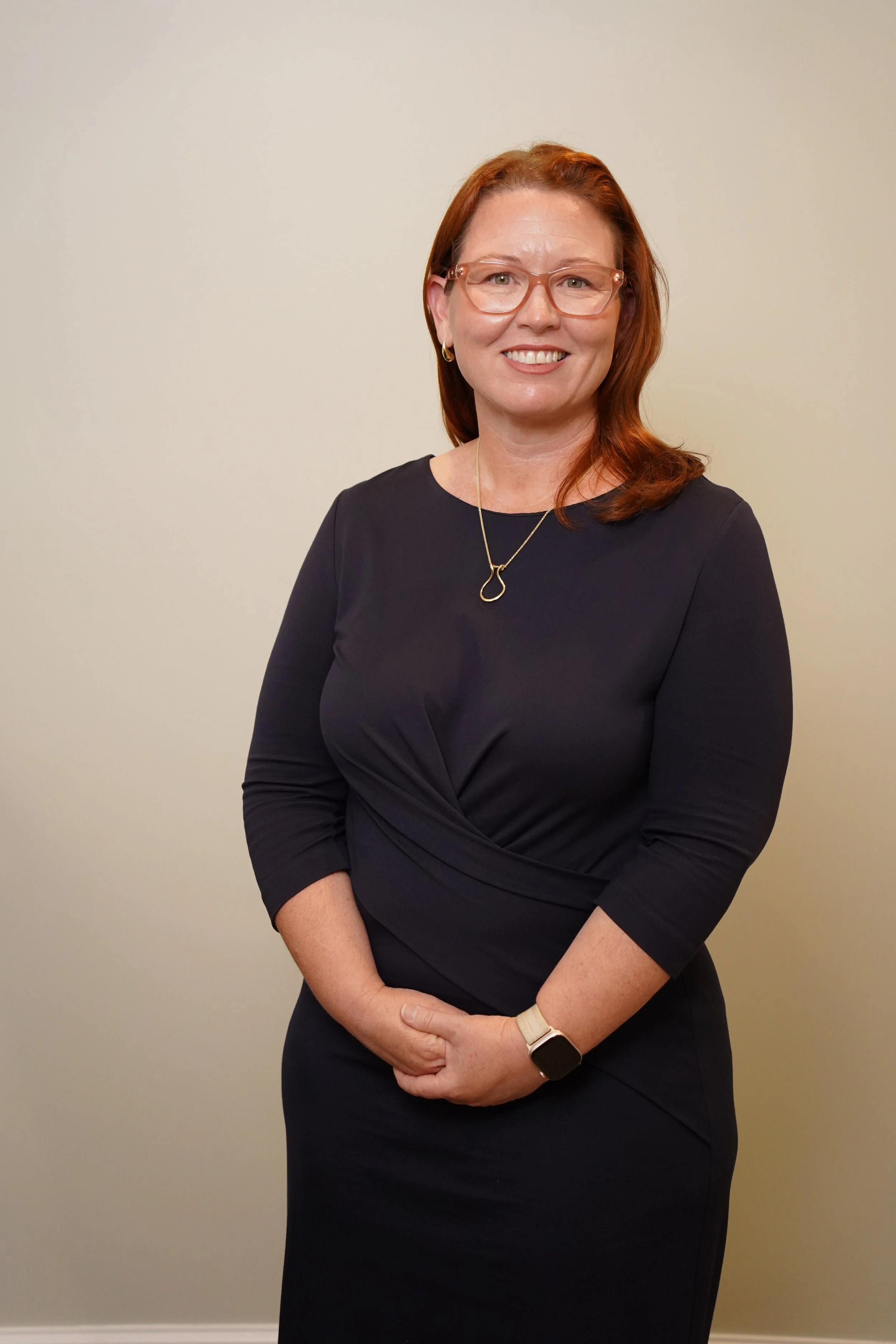 A woman with shoulder-length red hair, wearing glasses, a black dress, and jewelry, standing against a plain beige wall, smiling at the camera.