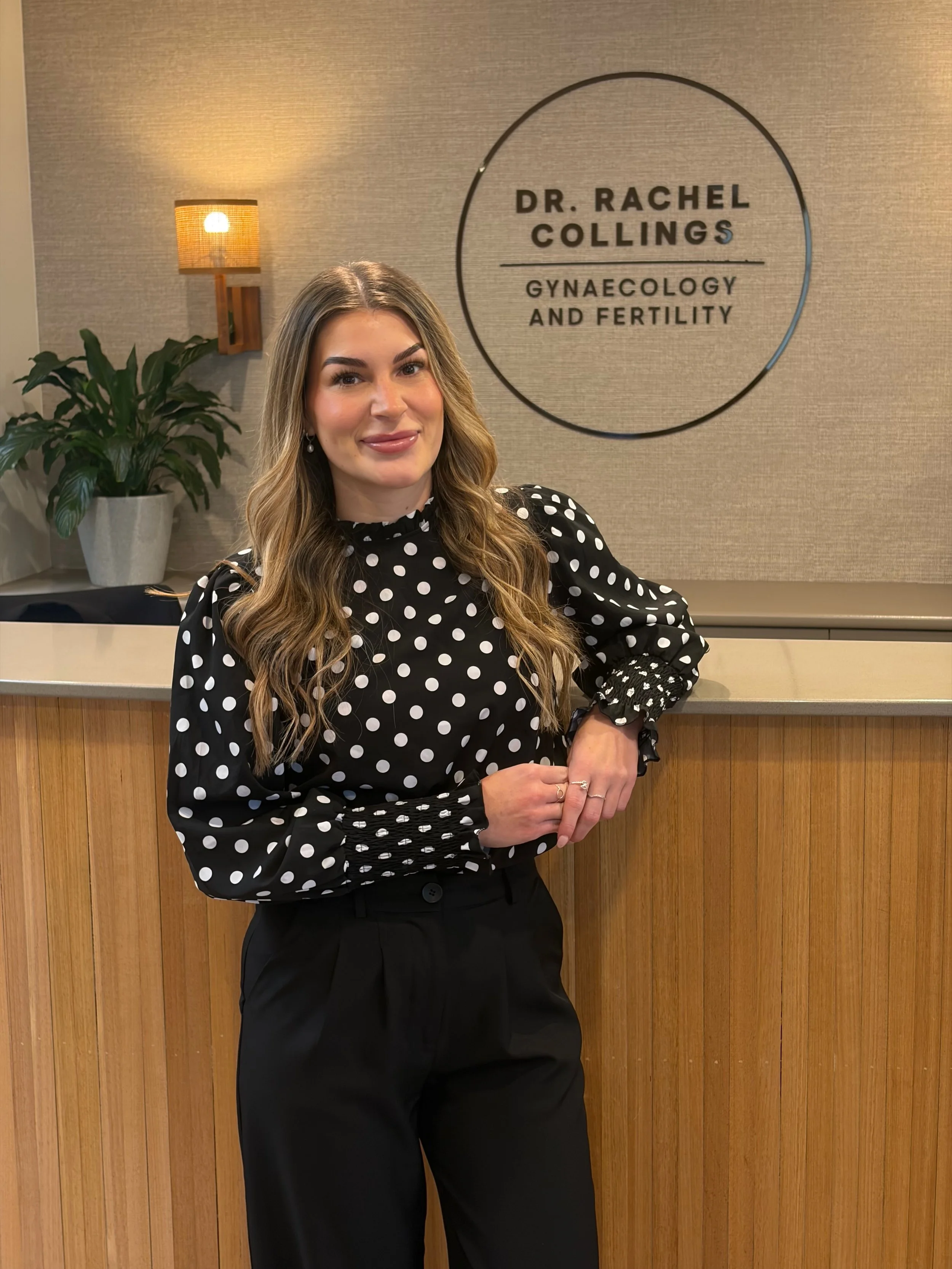 A woman with wavy blonde hair wearing a black polka dot long-sleeve blouse and black pants standing in front of a wooden panel wall. Behind her is a wall with a circular sign that reads "Dr. Rachel Collings Gynecology and Fertility". There is a potted plant and a wall-mounted lamp to her right.