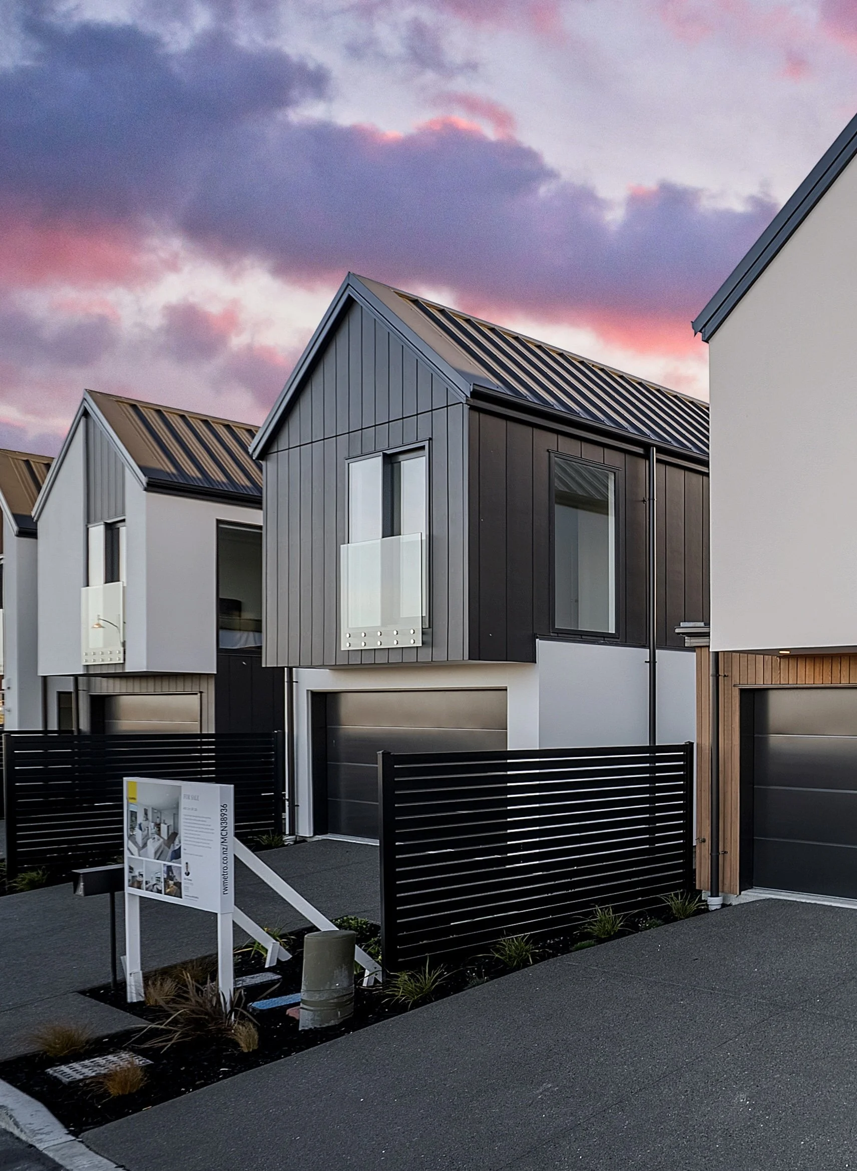 Modern townhouses with dark and white exteriors, large windows, and small front yards with trees and bushes, parked cars in driveways, under a partly cloudy sky.