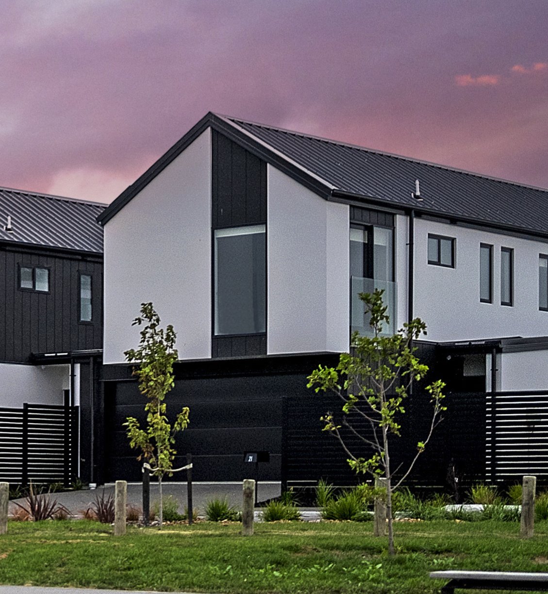 Modern row of three attached houses with different exterior colors, black garages, small front yards with trees, and a paved sidewalk under a blue sky with clouds.