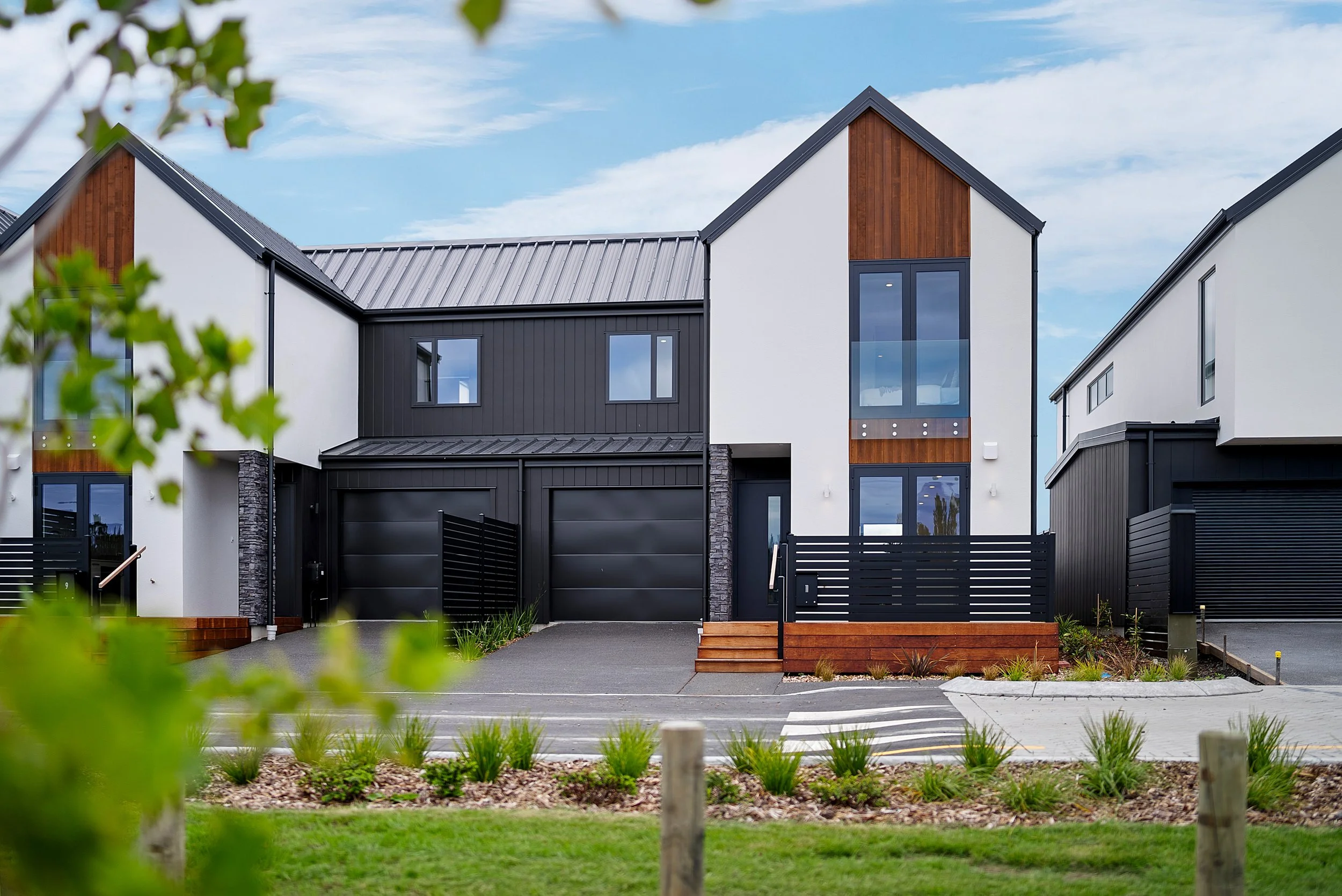Modern townhouse complex with multiple units featuring white and black exteriors with wood accents, black garage doors, front porches, and parking area with cars in front.