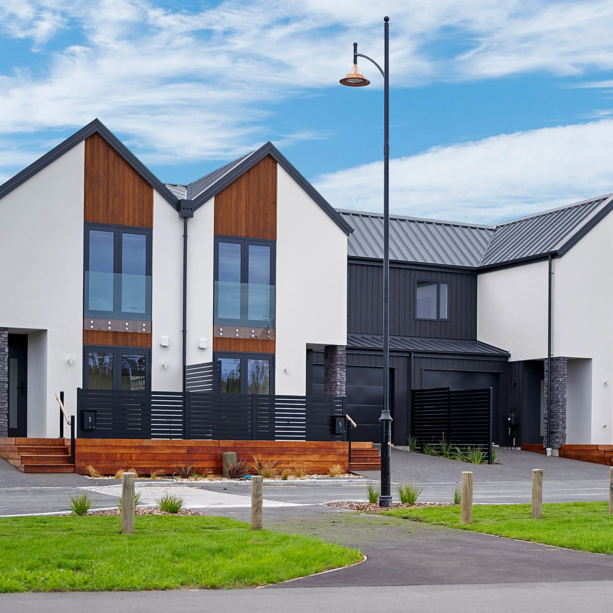 A row of modern townhouses with white and dark gray exteriors, gabled roofs, and front yards with greenery, cars parked in driveways, under a blue sky with scattered clouds.