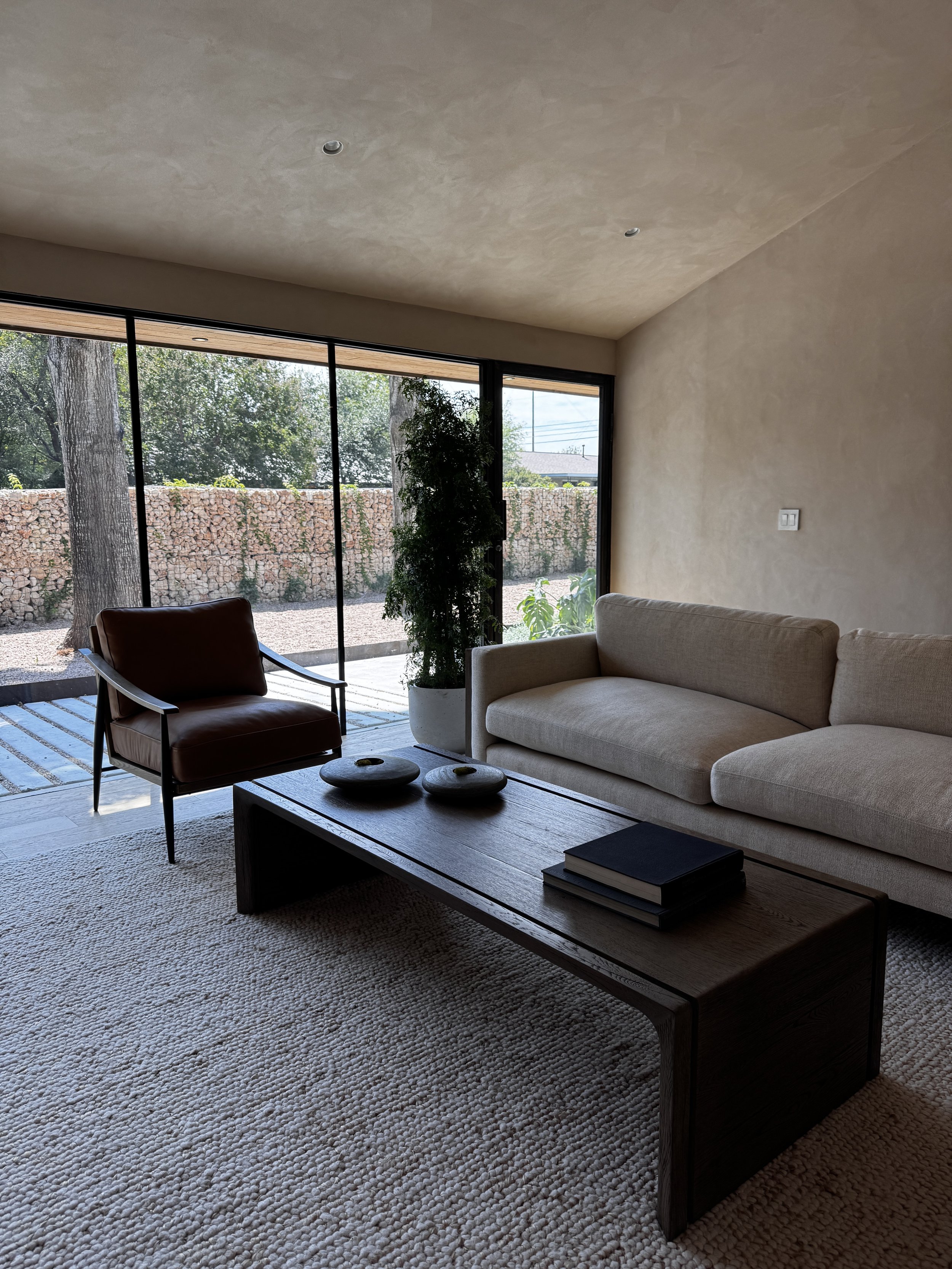 Living room with beige sofa, dark wood coffee table, brown armchair, large window with outdoor view of trees and stone wall, potted plant, and textured beige carpet.