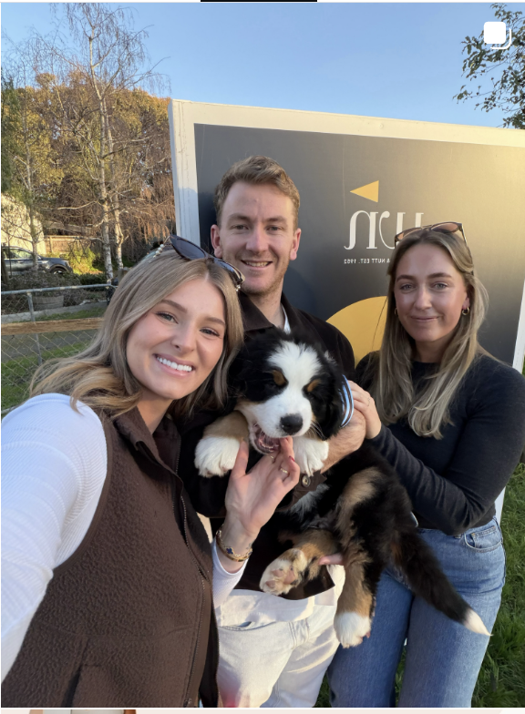 Three people taking a selfie outdoors with a black and white puppy, next to a sign with partially visible text.