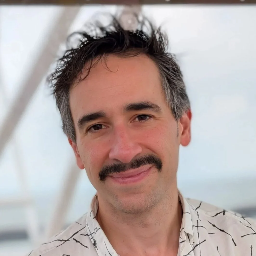 Portrait photo of David Labi in a white shirt, head angled, slight smile, with a cloudy sky in the background