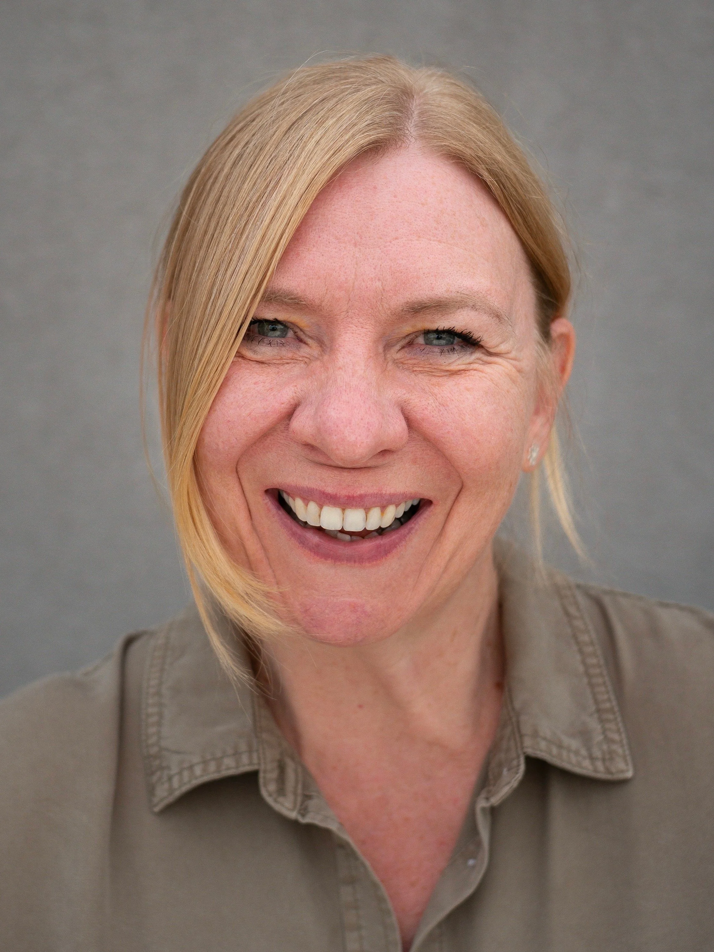 Porträt einer lächelnden Frau mit blonden Haaren, schwarzem Outfit und silberner Halskette vor einem weißen Hintergrund.