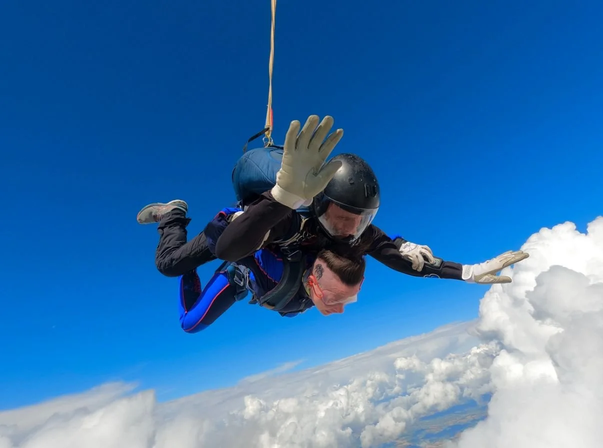 Two skydivers in freefall above the clouds, one in a black helmet and the other with a shaved head, both wearing jumpsuits and gloves, with a clear blue sky in the background.