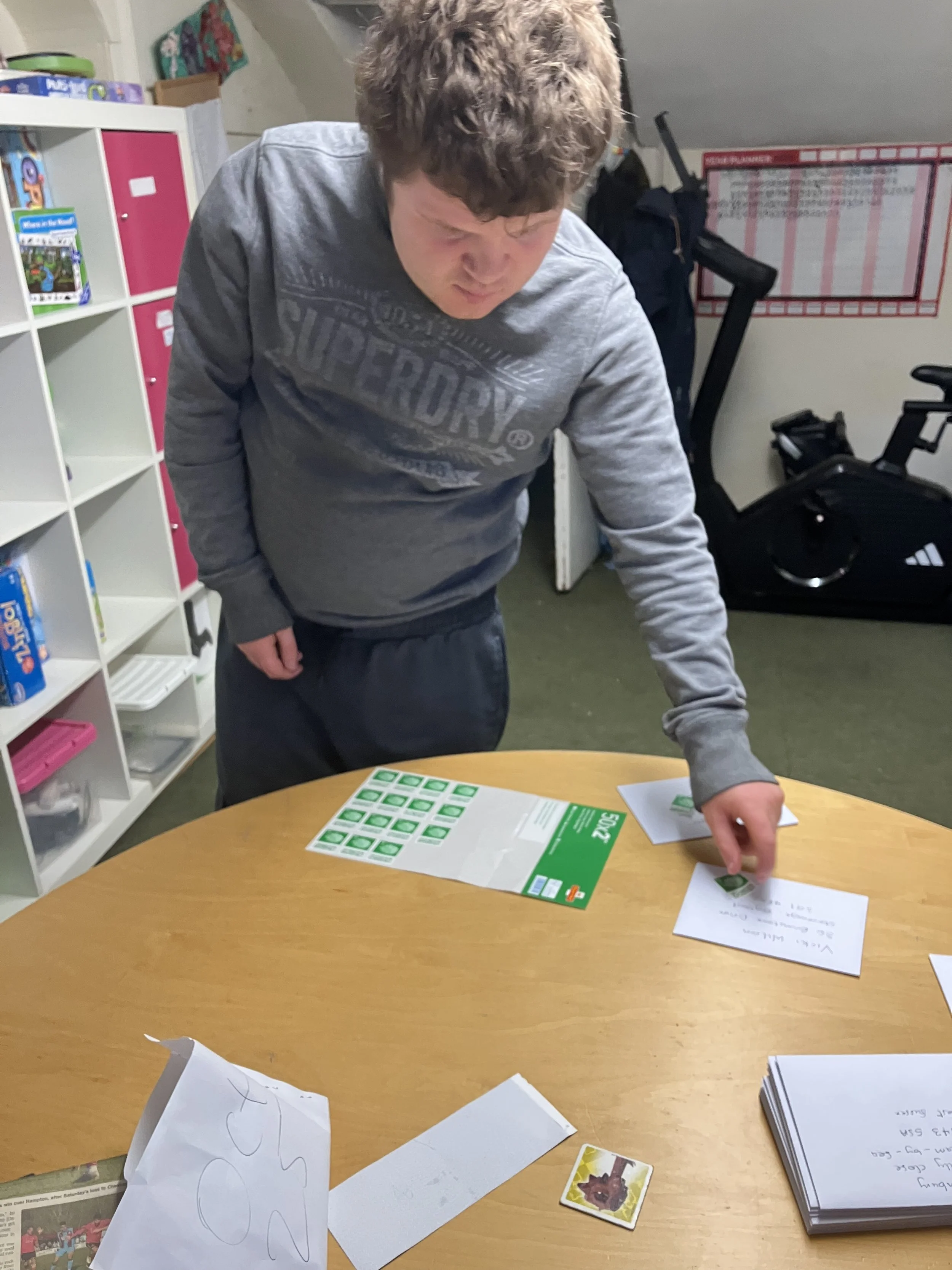 Young person with curly hair wearing a gray Superdry sweatshirt, standing at a wooden table, working with green stamps and papers.