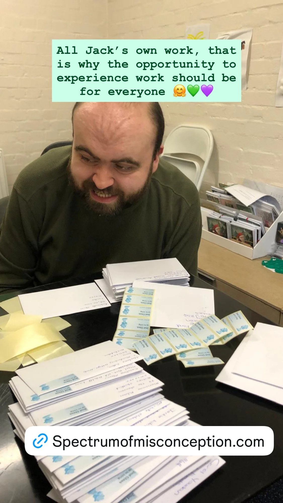 A man sitting at a table with stacks of envelopes and lottery tickets, smiling. There are papers on the wall and a box of more tickets or cards in the background.