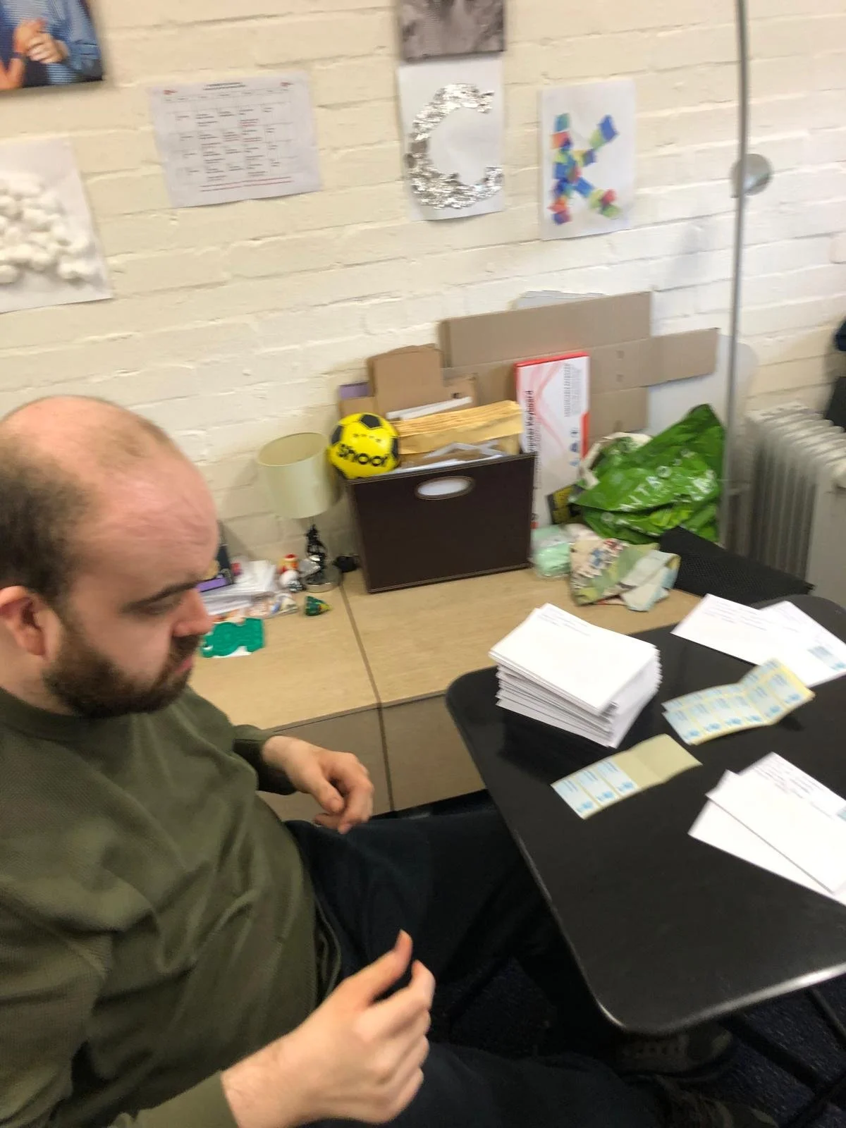 A man sitting at a desk with stacks of papers, envelopes, and a table with various items. The background has a white brick wall with artwork and decorations, including a yellow helmet, a silver decorative letter 'C', and paper crafts.