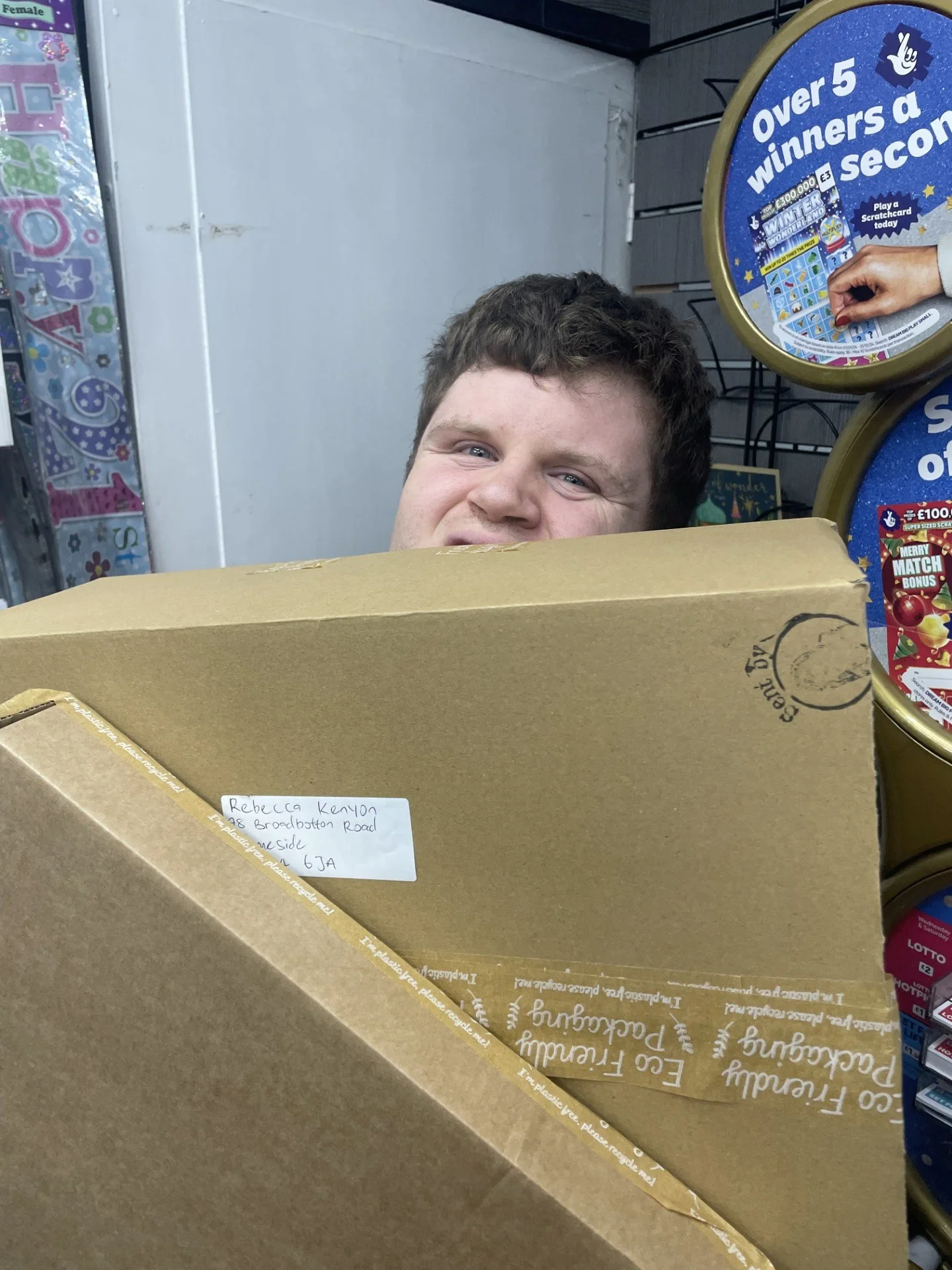 A young man is hiding behind a large cardboard box in a convenience store, peeking over it with a partial smile. The store shelves contain colorful lottery scratch-off tickets.
